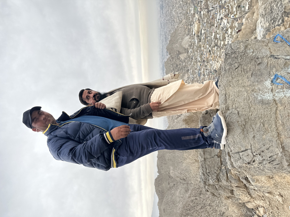 Two adult males are depicted standing on a prominent grey rock formation at a high elevation overlooking Quetta City Tehsil, Pakistan, under an overcast sky. The foreground rock features faint blue markings, including a '22'. The man on the left wears a black baseball cap, a dark blue quilted jacket with yellow and white trim, a blue t-shirt, dark blue athletic pants with a grey stripe, and blue and grey athletic shoes; he stands with his left foot forward and looks towards the camera. The man on the right, positioned slightly behind him, has a beard and wears a dark grey full-zip fleece jacket, a light beige shalwar kameez, and a light-colored patterned shawl draped over his left shoulder; he looks towards the right of the frame. The background reveals a dense urban landscape of Quetta City, characterized by numerous buildings and structures, nestled amongst barren, rocky hills that extend to the horizon.