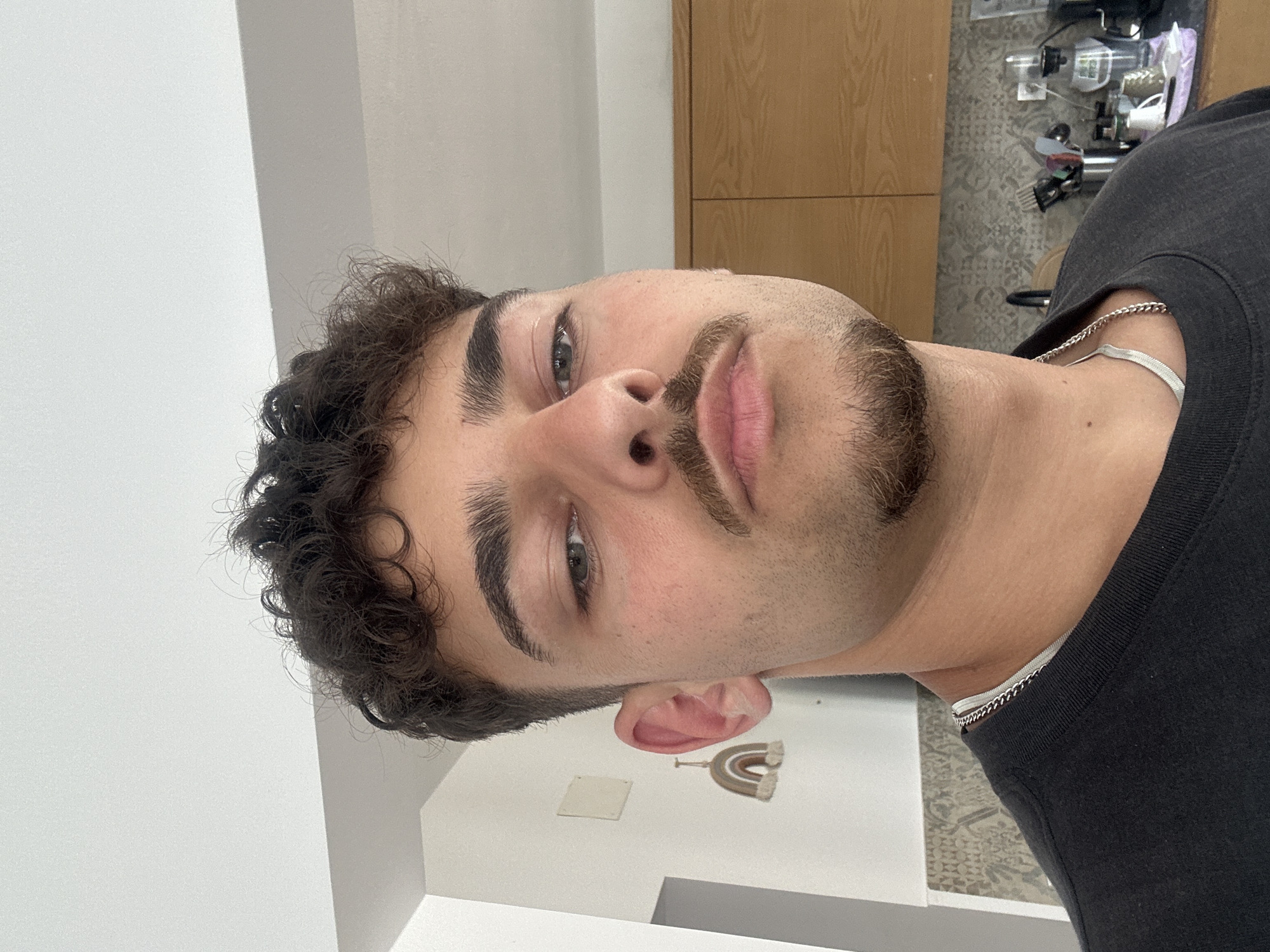 A close-up, angled portrait of a young man with curly dark hair, light skin, and a trimmed mustache and beard. He wears a black t-shirt and a silver necklace. The background includes a white wall, light brown wooden cabinets, and a patterned floor. A small decorative rainbow hangs on the wall. The location in the city of Troyan, Bulgaria cannot be determined from the image.