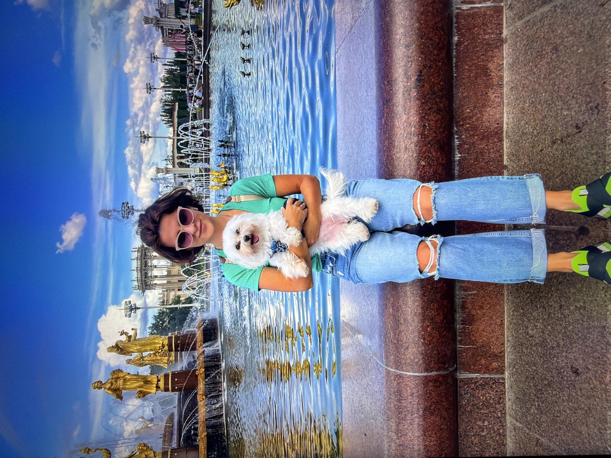 A woman with short brown hair and sunglasses is holding a small white dog. She is wearing a green t-shirt and ripped blue jeans. She is sitting on a stone ledge in front of a large fountain with golden statues. The fountain is surrounded by water, and in the background, there are more fountains and buildings. The sky is blue with some white clouds. The location is the Friendship of Peoples fountain in the city of Kotelniki, Russia.