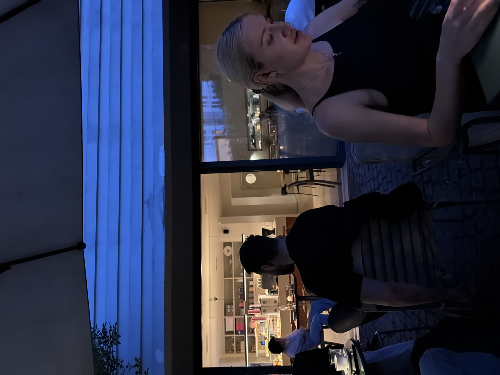 The image depicts two people sitting at an outdoor cafe in Belgrade, Serbia. A blonde woman is in the foreground, wearing a black tank top and silver necklace, with her eyes closed. A man with a beard and black shirt sits behind her, both seated at chairs in a cafe setting, visible through a large open doorway. The cafe's interior features shelves of desserts and a service counter. Outside, blue slat panels provide a backdrop, with a partial view of an umbrella. The overall lighting suggests evening.