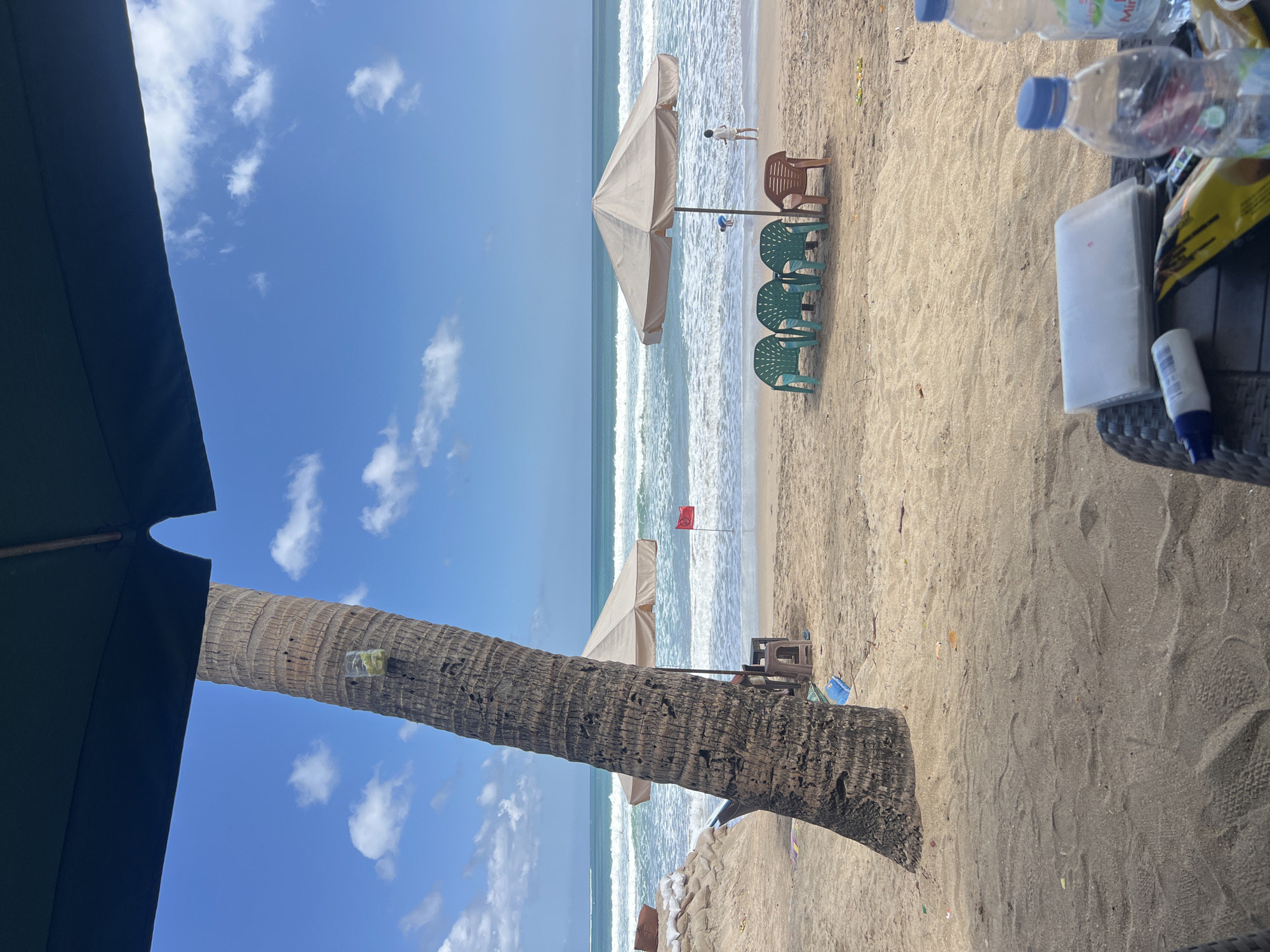 A view from beneath a large dark green umbrella on a sandy beach in Kuta, Indonesia. A thick palm tree trunk is to the left, with a small plastic cup containing what appear to be yellow slices, possibly lemons, attached to it. In the mid-ground, the ocean with white waves stretches to the horizon under a bright blue sky with scattered white clouds. Two beige beach umbrellas are set up near the water's edge, one with a person sitting under it and a small red flag on a pole nearby. A group of four plastic chairs, three green and one brown, are arranged in front of one of the umbrellas. Further down the beach, a child is visible near the water. The foreground shows a sandy area with a black woven table holding a stack of white papers in a clear plastic sleeve, a small white bottle, and two plastic water bottles, one of which has a blue cap and a label that reads "Mir..." indicating it is mineral water. Some debris, including a yellow and black bag, is scattered on the sand. The scene appears to be a typical beach resort setting.