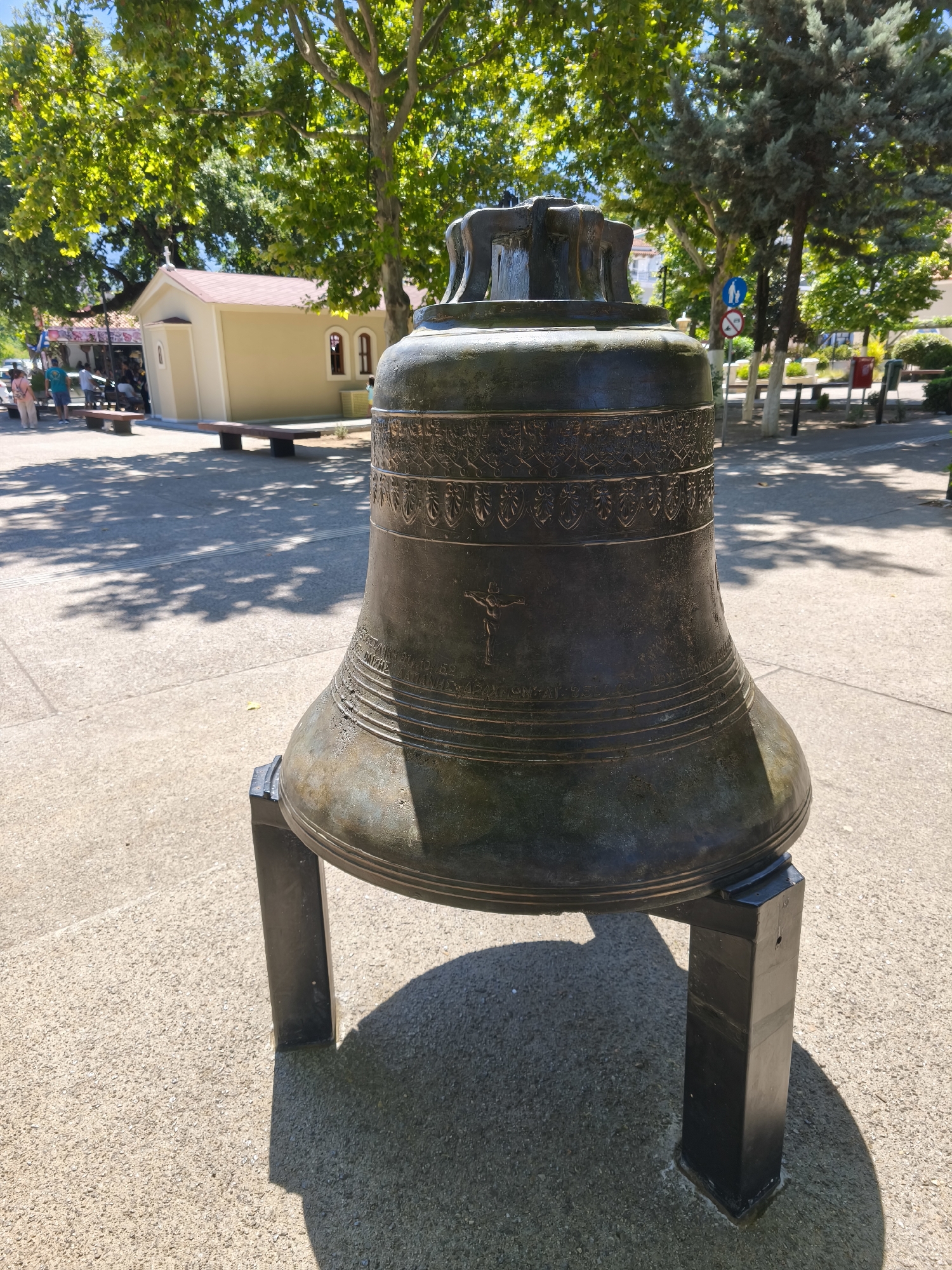 A large, dark bronze bell rests on a black metal stand in the foreground of a sun-dappled outdoor area. The bell is intricately decorated with bands of ornamental patterns and an image of the crucifixion. Behind the bell, a small, light-colored building with a red roof, possibly a chapel, is visible, with a few people and park benches in the vicinity. The ground is paved with concrete, and the scene is framed by lush green trees. The overall impression is a quiet, possibly religious, outdoor setting in the town of Prokopi, Greece.