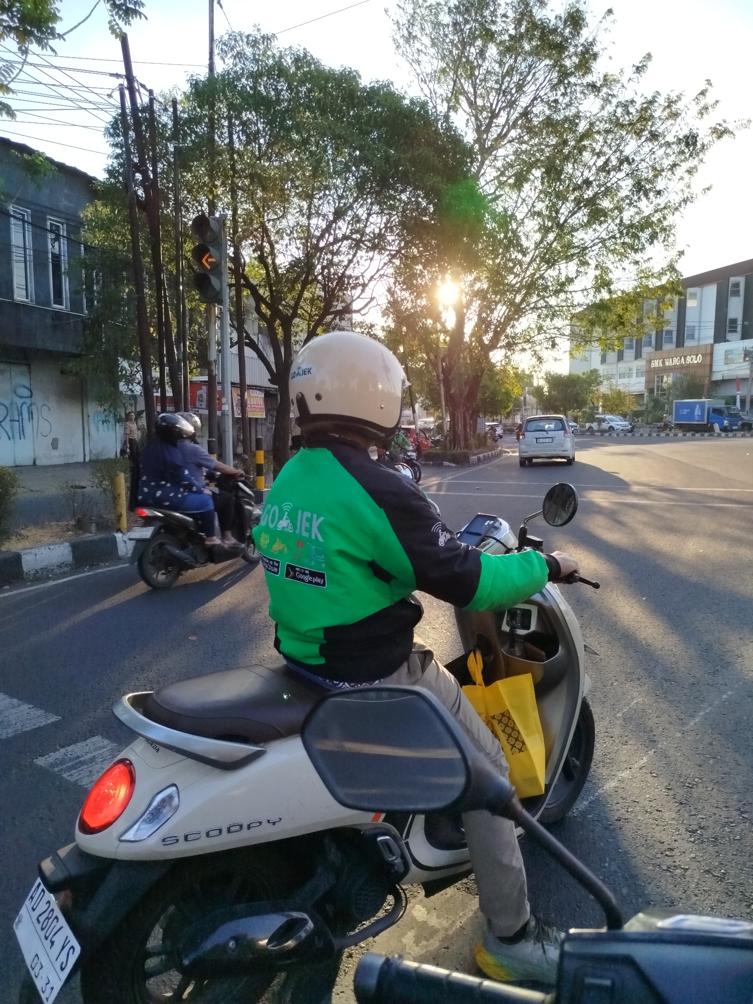 In Surakarta, Indonesia, on a street bathed in the warm glow of late afternoon sun, a Gojek driver, clad in a distinctive green jacket and white helmet, is stopped at a traffic light. The driver is seated on a white Honda Scoopy scooter, with a yellow shopping bag tucked into the front storage compartment. The scooter's brake light is illuminated, indicating it is either stopped or slowing down. Another scooter carrying two passengers is visible in the left background, also stopped at the light. The traffic signal shows a flashing yellow arrow, suggesting caution. Buildings line the street, one of which is labeled "SMK WARGA SOLO." The road surface is asphalt, with white pedestrian crossing lines visible in the foreground. The scene captures a moment of urban transit in Surakarta, where Gojek services are a common sight.