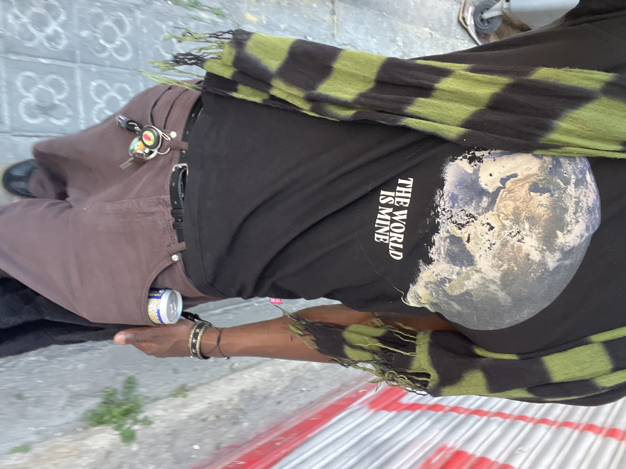 The image, taken in Barcelona, Spain, captures a close-up, angled view of a person outdoors during the daytime. The individual, whose face is not visible, is wearing a black short-sleeved t-shirt, brown pants, and a green and black striped scarf with fringed ends draped over their shoulder and arm. A black belt with a silver buckle cinches the pants, and a set of keys with a small flashlight and colorful keychain is clipped to a belt loop. On the person's right wrist, multiple bracelets are visible, including beaded and metallic designs.
A silver and blue beverage can is tucked into the side pocket of the brown pants. The black t-shirt features a large graphic of the Earth (planet) and vertical white text that reads, "THE WORLD IS MINE."
The setting appears to be a street or sidewalk. The ground consists of distinctive grey pavement tiles, some of which exhibit a common Barcelona 'panot' pattern, and also features red and white painted lines or stripes in the lower right. Small patches of green vegetation grow from cracks in the pavement. The lighting suggests it is daytime.