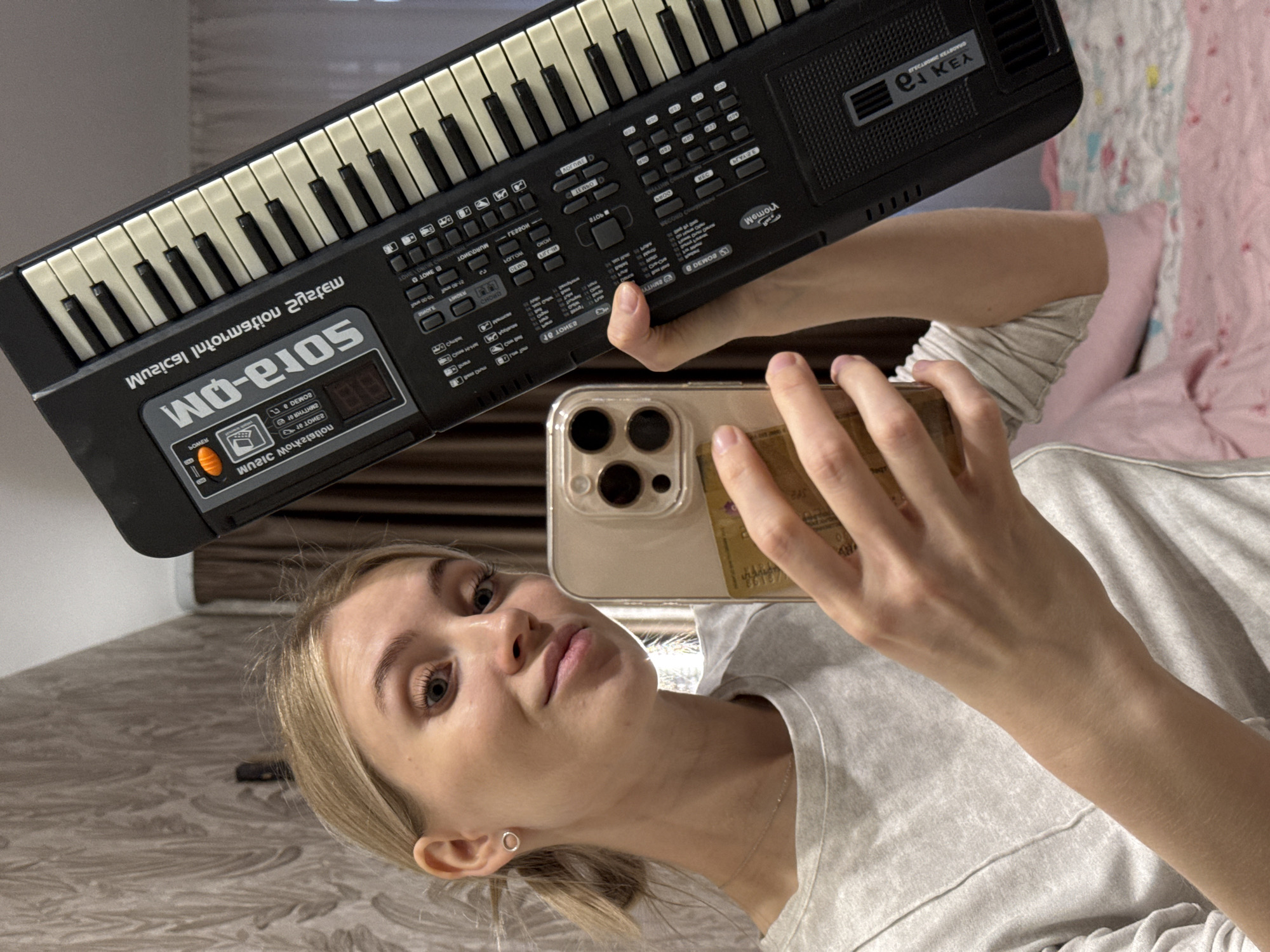 A blonde woman is holding a black and white electronic keyboard at an angle while taking a selfie with her smartphone. The keyboard is labeled "Sora-OM WO-210S." The woman is wearing a light gray shirt and a small earring. The background includes a glimpse of a bed with pink bedding. There's no indication of the location being Izhevsk, Russia.