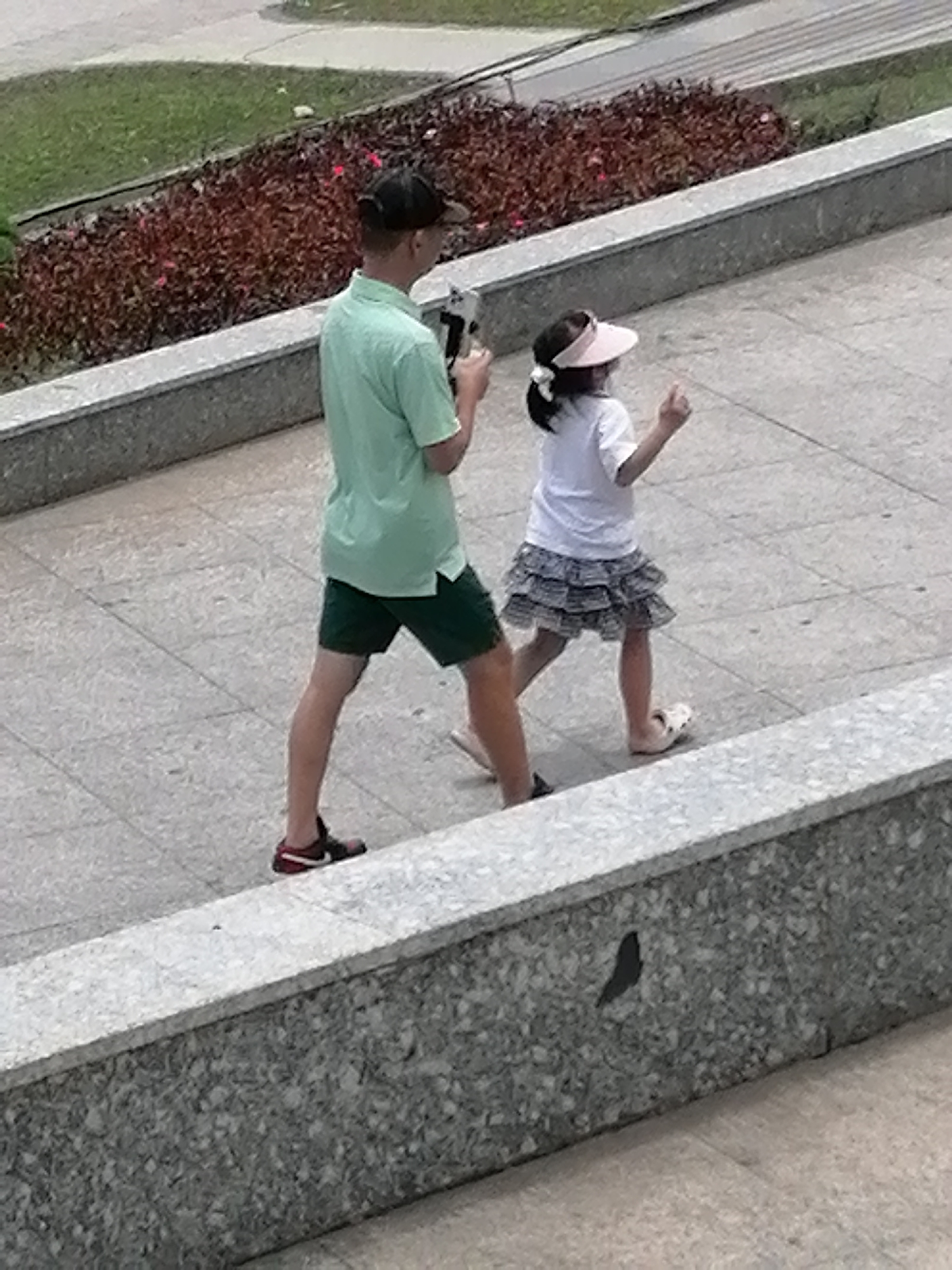 In Da Lat, Vietnam, a man and a young girl are walking on a paved path. The man, wearing a light green t-shirt, dark green shorts, and a black baseball cap, is holding what appears to be a camera or phone in his right hand. The girl, dressed in a white t-shirt, a layered grey and white skirt, and a pink visor, has her left hand raised, possibly gesturing or walking. They are positioned on a tiled pathway next to a raised planter bed filled with reddish-brown foliage and some small pink flowers. The pathway is bordered by a grey granite-like structure. The overall environment suggests an outdoor public space, potentially a park or a promenade in Da Lat.