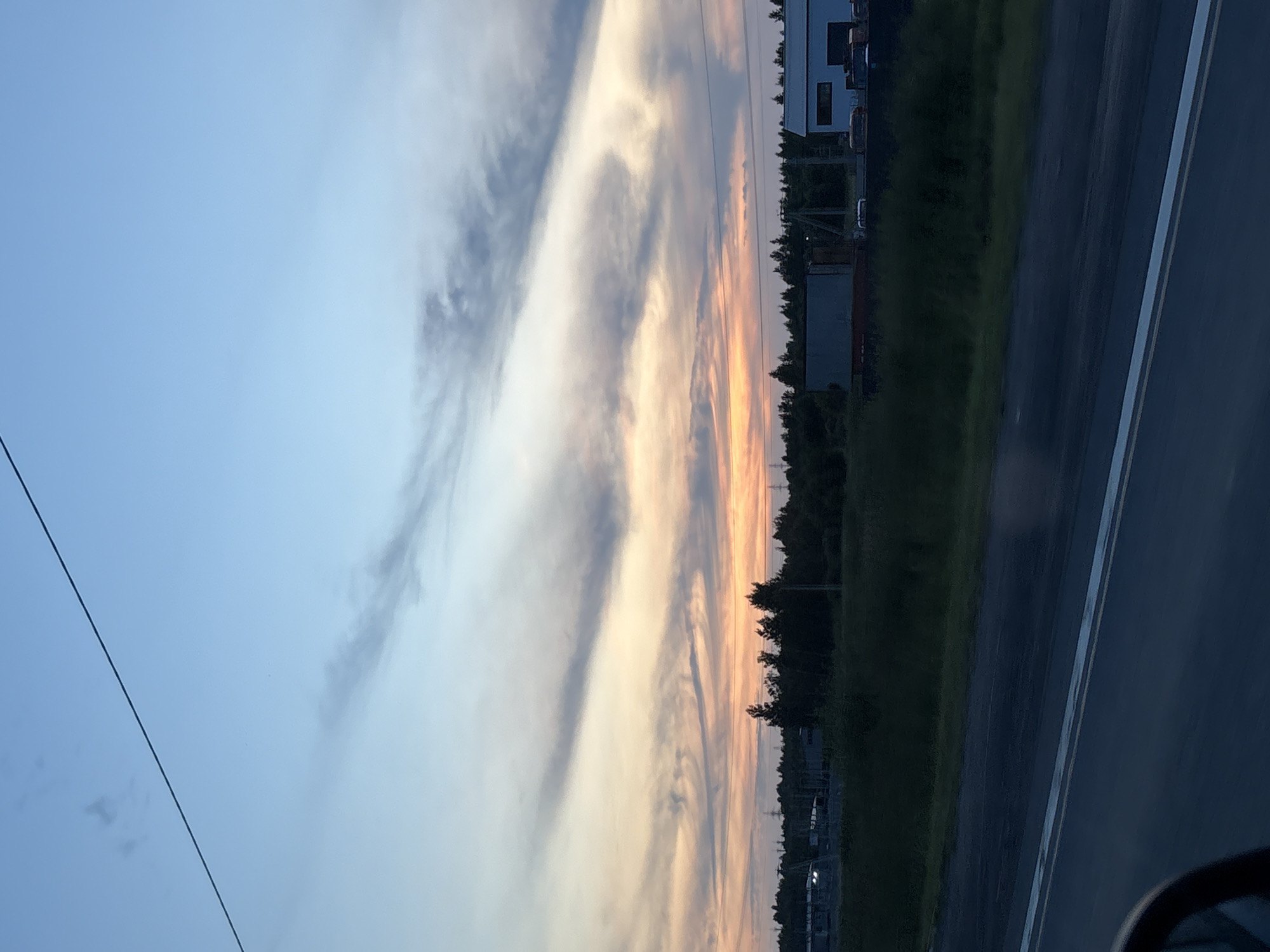 The image shows a wide shot of a twilight sky over a rural landscape. The sky is a gradient of blue, fading into soft oranges and pinks near the horizon, with wispy clouds streaking across. A thin black wire stretches diagonally across the upper portion of the frame. Below the sky, a dark line of trees separates the sky from the foreground. Beyond the trees, there are a few industrial-looking buildings and structures, some with lights on. The foreground consists of a dark, paved road with white markings, suggesting it's a highway or a main road. The edge of a vehicle's side mirror is visible in the bottom left corner, indicating the photo was taken from a car. The scene appears to be a view from a moving vehicle passing through a semi-rural area near Ozerki, Russia, at dusk.