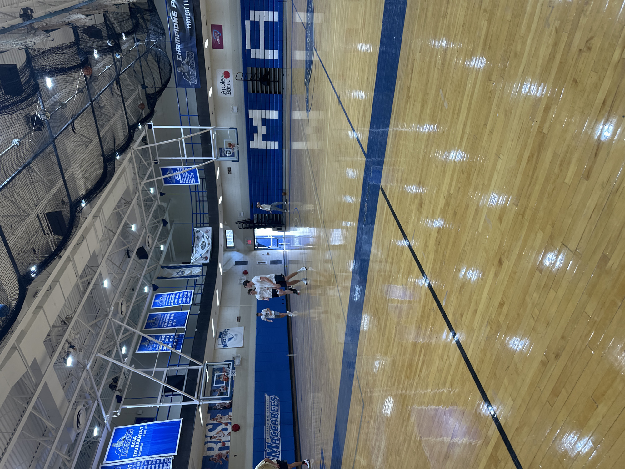 This vertical indoor shot captures a basketball gymnasium, likely at Yeshiva University in New York, USA. The wooden court, with its shiny surface reflecting the overhead lights, features prominent blue lines and a large blue section beneath the far basket. Several young men, appearing to be players or students, are scattered across the court, some in white t-shirts and shorts/pants, with one individual visible near the foreground. Two basketballs are on the court.
The gym features multiple basketball hoops and blue bleachers along the far wall, where large white letters, possibly forming "YUH" or "YM," are visible. The ceiling is high, with exposed white beams and extensive black protective netting. Numerous blue and white banners hang from the ceiling structure, commemorating various achievements of the "Yeshiva University Maccabees" basketball team, including "NCAA Tournament" appearances (e.g., 2014, 2015) and "CHAMPIONS" titles (e.g., 1987, 1999, 2017, 2017-2018, 2019-2020, 2021-2022, 2024-2025). Other banners read "MACCABEES UNITE" and "PROTECT THE" (partially visible). An "Apple Bank" sponsorship logo is also visible on the wall. The scene suggests a practice session or casual activity within the university's sports facility.