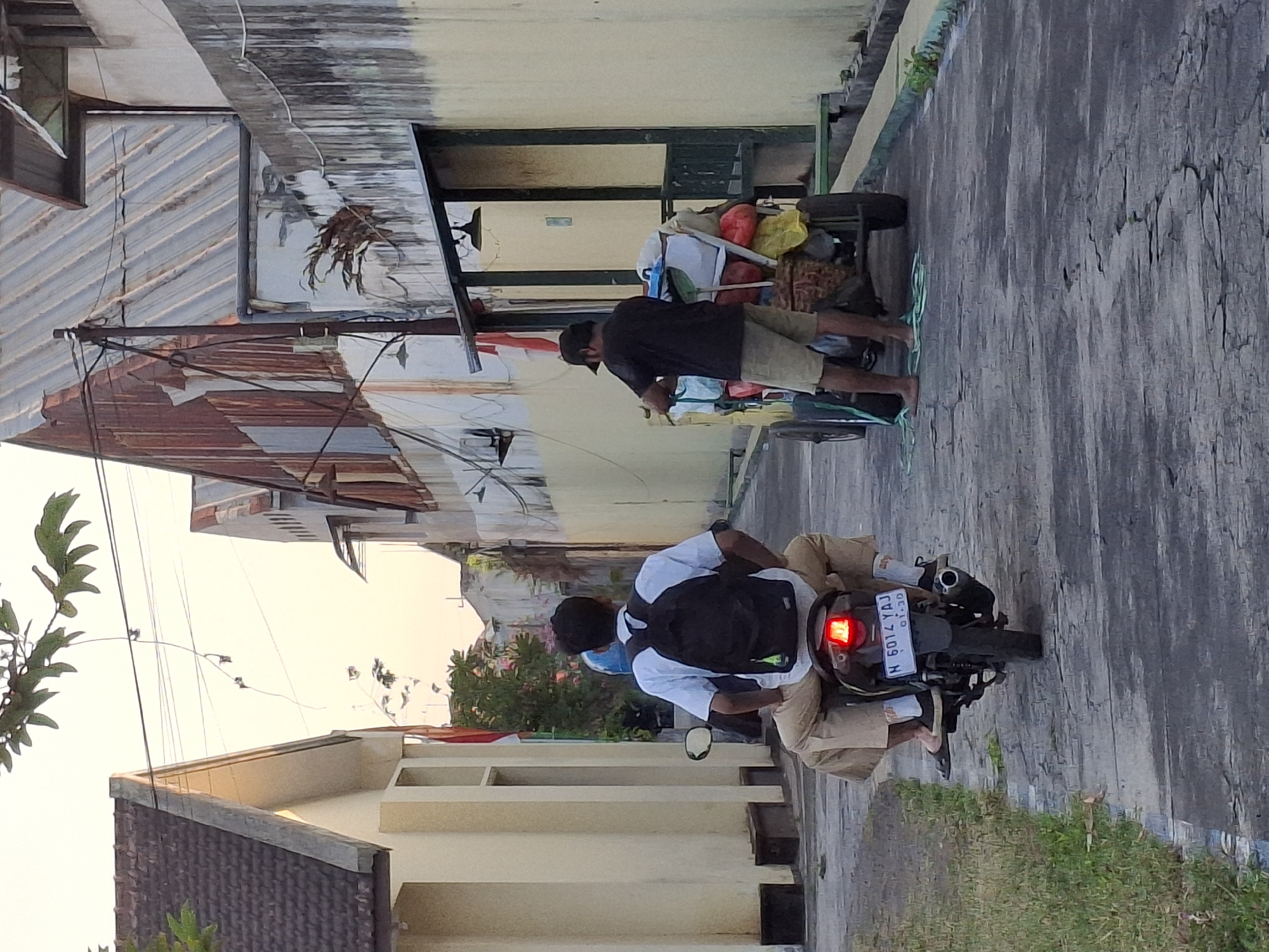The image depicts a narrow street in Lumajang, Indonesia, likely in an industrial or commercial area, characterized by buildings with weathered and rusted metal roofs alongside lighter-colored structures. In the foreground, a person is riding a motorcycle, wearing a white shirt and dark backpack, with their legs extending to the sides. The motorcycle's license plate reads "N 6014 YAJ". Further down the street, another person, wearing a black t-shirt and khaki shorts, is standing behind a wheeled cart laden with various items, including bags and what appear to be food items wrapped in plastic. The cart is positioned on the right side of the street. A flagpole with an Indonesian flag is visible on the left side of the street, attached to one of the buildings. The sky suggests it is late afternoon or early evening, with soft lighting. The ground is paved with a textured surface that appears worn and cracked in places, with a strip of grass bordering the left side of the street.