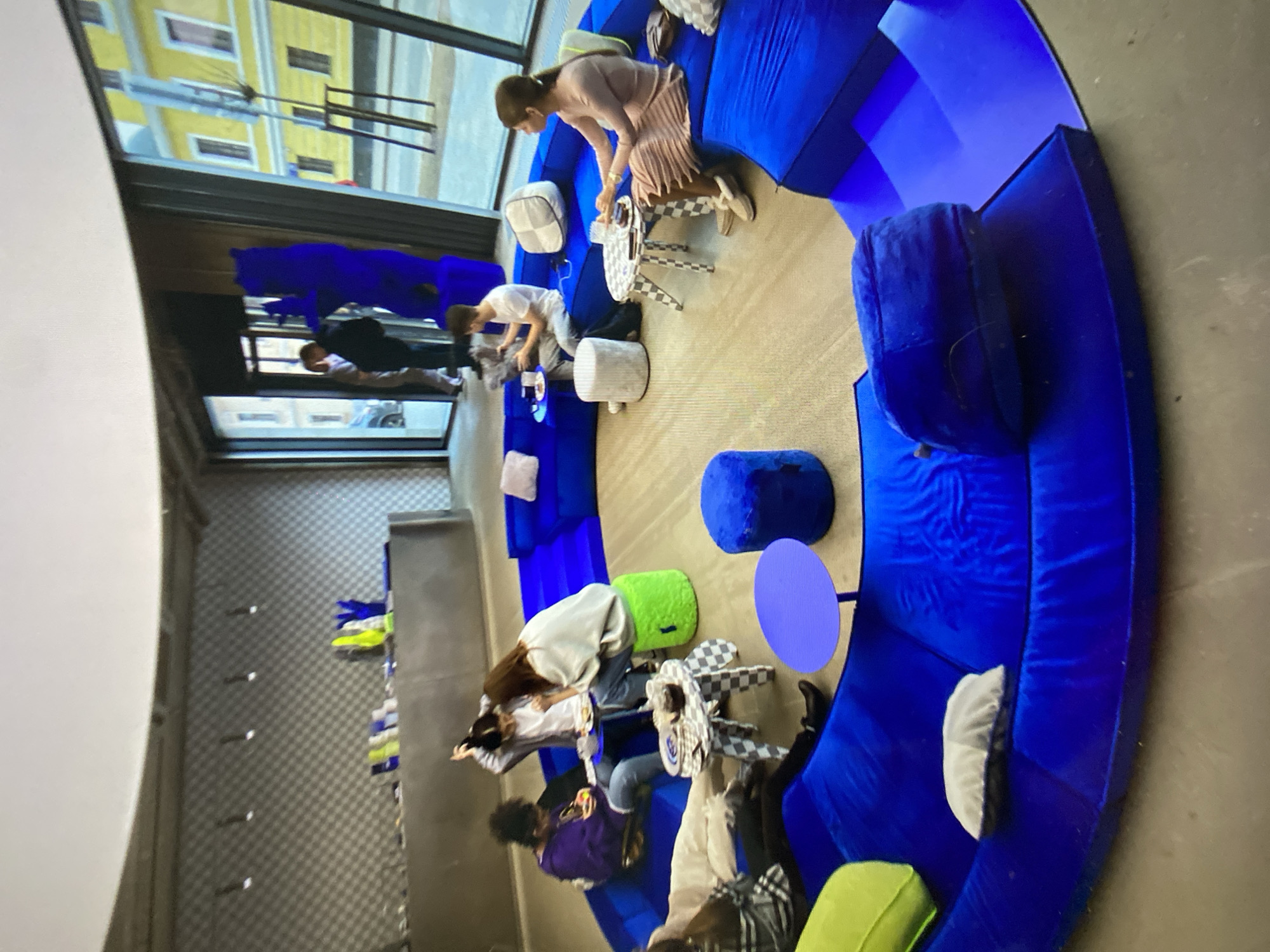In a modern, open-plan interior, likely a retail or communal space, a large, circular, sapphire-blue modular sofa dominates the scene. Several individuals are seated on the sofa, engaged in various activities. To the left, a person in a purple hoodie and jeans is looking at their phone. Next to them, two individuals are seated, one in a grey hoodie and the other in a white sweatshirt, appearing to be interacting over a small checkered table with food and drinks. Further around the curve of the sofa, a person in a white t-shirt is seated, holding an object. To their right, a person in a pale pink dress is seated, holding a glass. Various circular tables with checkered patterns and small poufs are scattered around the seating area, some with drinks and snacks. The environment features a patterned wall in the background with lighting fixtures, and large windows that offer a view of a yellow building facade and streetscape, suggestive of an urban setting in Khimki, Russia. The overall atmosphere is relaxed and contemporary.