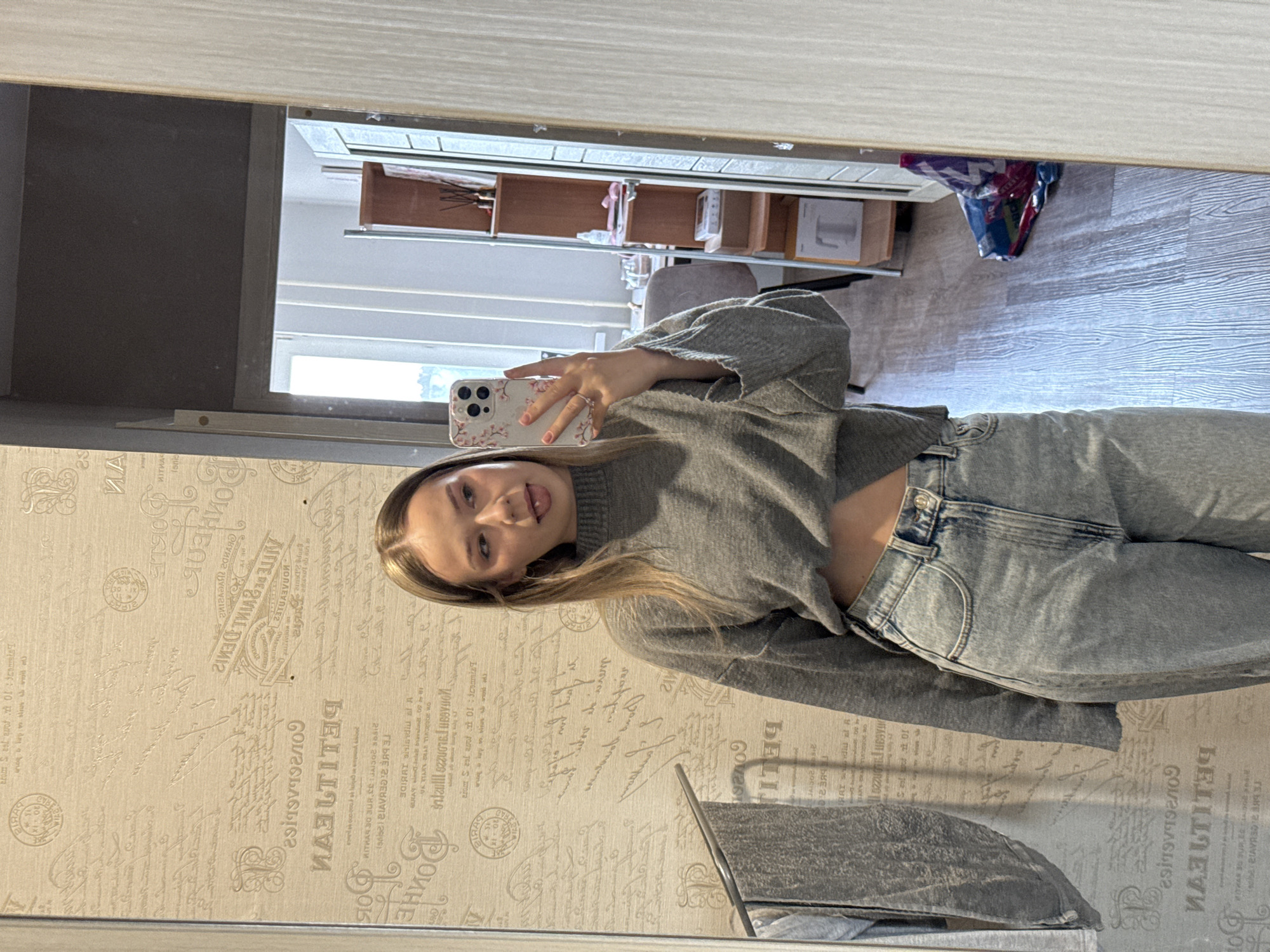 A young woman with blonde hair is taking a selfie in a mirror. She's wearing a gray sweater that exposes her midriff and light blue jeans. The mirror reflects a room with a light wood floor and a shelf with various items on it. The wall behind the mirror has a patterned wallpaper with text and decorative elements. The OCR text is mirrored. There is a metal towel rack. The location is indicated in the mirrored text on the wallpaper and the city of Chaikovsky, Russia is not indicated in any way in the image.