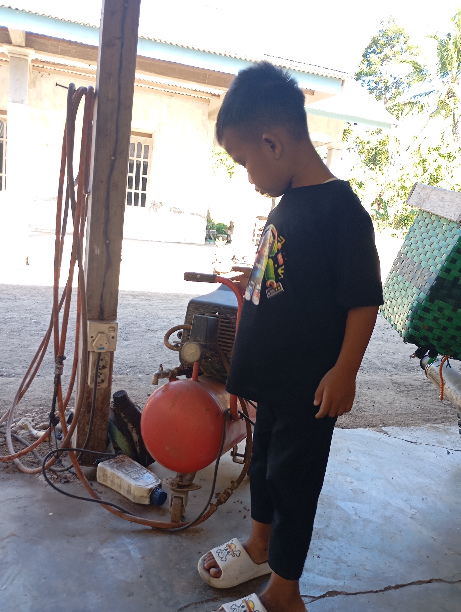 In Ketepung, Indonesia, a young boy stands in a dusty outdoor area, his gaze directed downwards. He is wearing a black t-shirt with a colorful graphic and black pants. His feet are clad in white sandals with cartoon characters. To his left, a wooden post supports several thick, coiled orange hoses, a white electrical outlet with multiple sockets, and a black cable. Behind the boy and to his right, a red air compressor with a gauge and a spigot is visible. A white plastic bottle with a blue cap lies on the ground near the compressor. In the background, a light-colored building with visible window frames stands under a blue-trimmed roof. The ground is a mix of dirt and concrete. The scene is illuminated by bright daylight, casting distinct shadows.