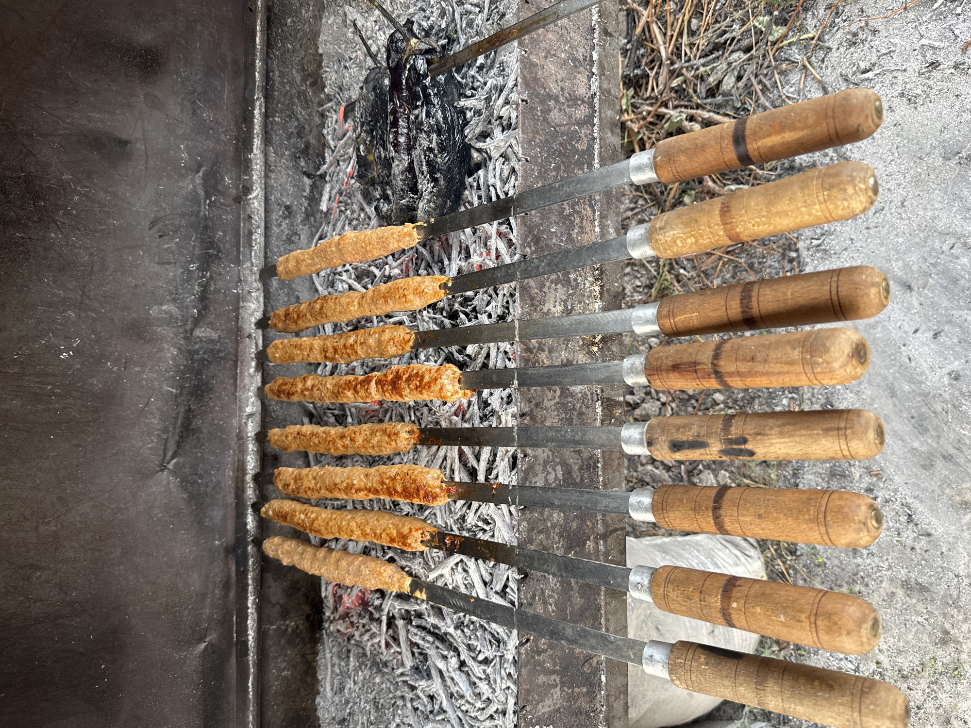In Aghavnadzor, Armenia, a barbecue grill holds ten skewers of grilled meat. The skewers are arranged horizontally above a bed of ash and glowing embers. The meat on the skewers is elongated and appears to be ground meat, possibly lamb or beef, cooked to a light brown color with some charring. The handles of the skewers are made of wood. The grill itself is made of metal and shows signs of use. The background is out of focus, but appears to be an outdoor setting with dry vegetation and loose soil.