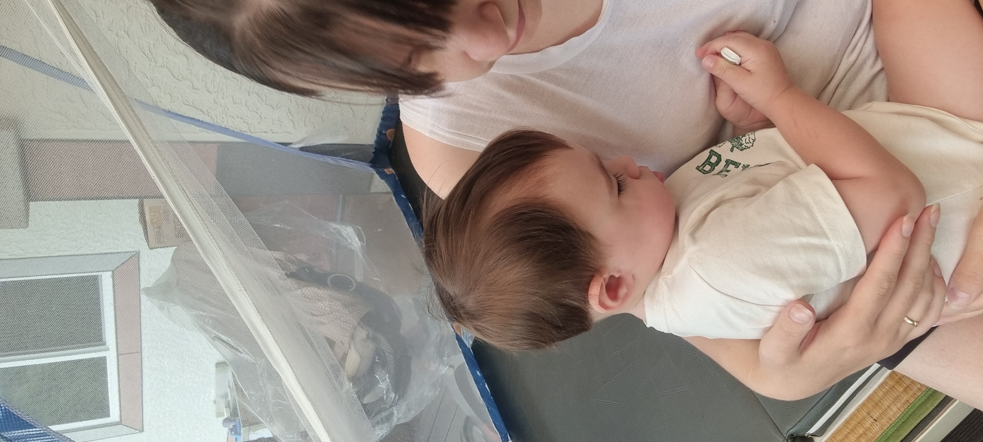 An intimate, high-angle photograph captures a mother holding her baby or toddler indoors. The mother, with short, dark hair, is visible from the chest up, wearing a plain white t-shirt. She looks down tenderly at the child cradled in her arms. A gold ring is visible on her left hand.
The baby, likely an infant or young toddler, has light brown hair and is sleeping peacefully with closed eyes and a slightly parted mouth. They wear a cream-colored t-shirt with green text on the chest, partially visible as "BEAR" or a similar word. The child's left hand is holding a small, white, cylindrical object.
The scene is set indoors during the daytime, with natural light illuminating the subjects. A sheer white mesh curtain or netting partially covers the left side of the frame, possibly part of a playpen or a protected sleeping area. Through the mesh, a textured wall and a white-framed window with multiple panes are visible in the background. Beneath the child, a dark grey or black surface is present, and to the lower right, a textured, woven mat, resembling a tatami mat, is visible. The background also reveals a glimpse of a child's area behind the mesh, containing a black object and other items under a transparent plastic sheet.
The overall atmosphere is one of peaceful rest and maternal affection. The location is Dunnae-myeon, South Korea.
