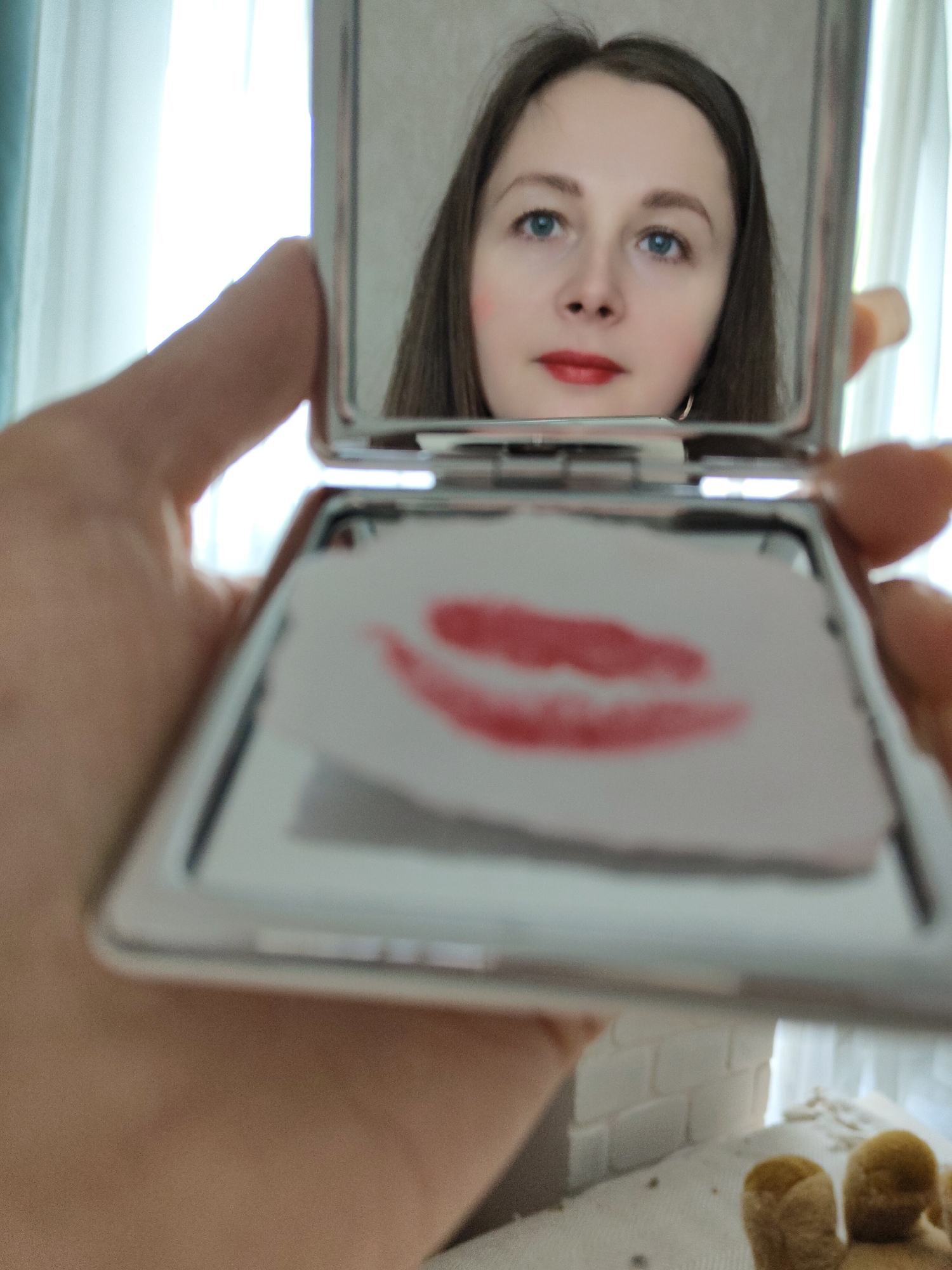 A woman is visible in the reflection of a silver compact mirror held in a hand. The compact is open, revealing a paper with a lipstick kiss imprint on it. The woman in the reflection has light skin, blue eyes, brown hair, and is wearing red lipstick. In the background, there are white curtains. The location is Unknown, Unknown.