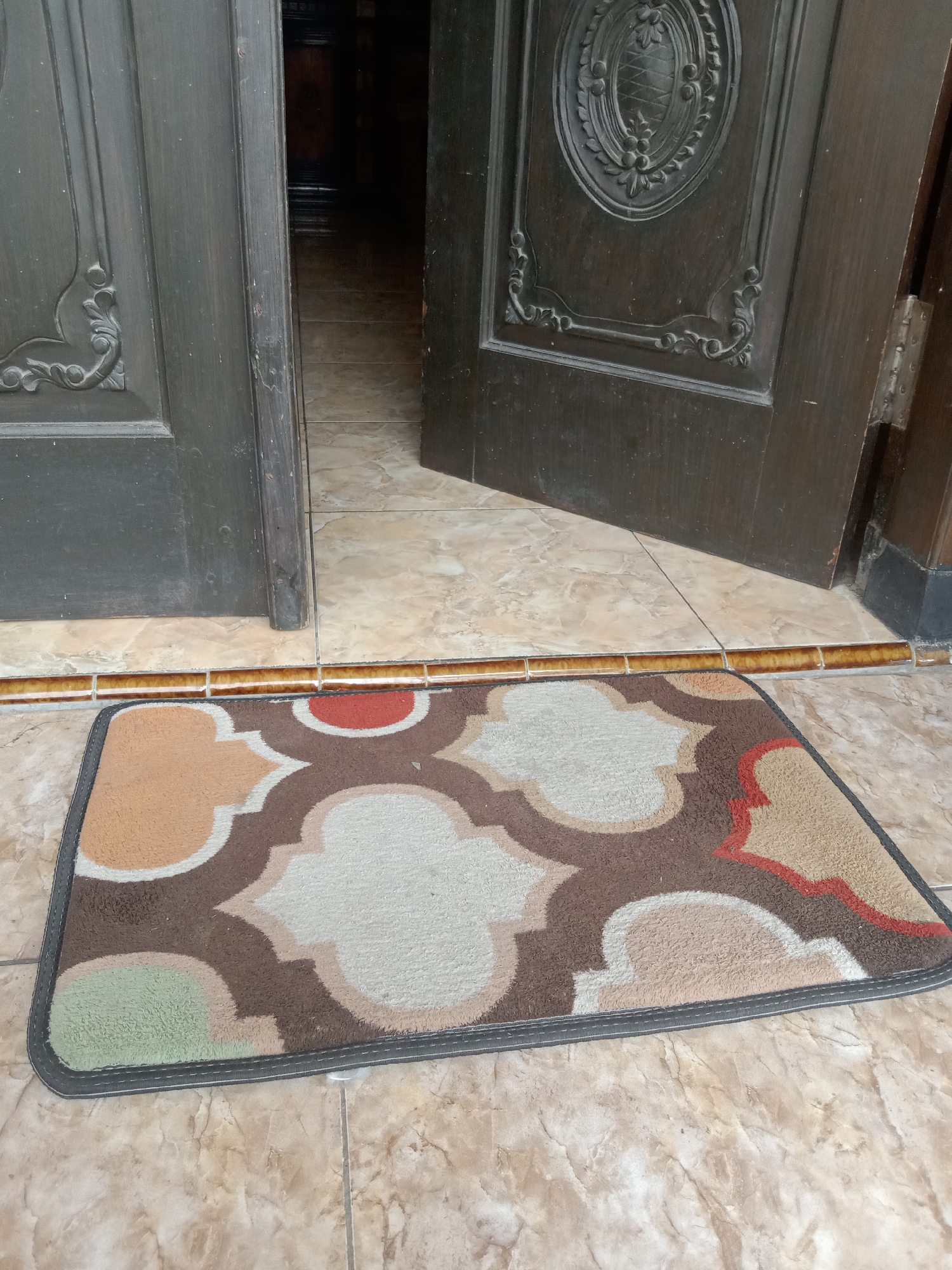 A doormat with a patterned design featuring brown, tan, white, red, and green shapes is placed on a tiled floor in front of an open dark wooden door. The door has ornate carvings on its surface. The floor tiles are beige with a marbled pattern. The doormat is positioned at the threshold of the doorway, leading into a dimly lit interior. This scene is likely a doorway in a residential or public building in Stiaji, Indonesia.