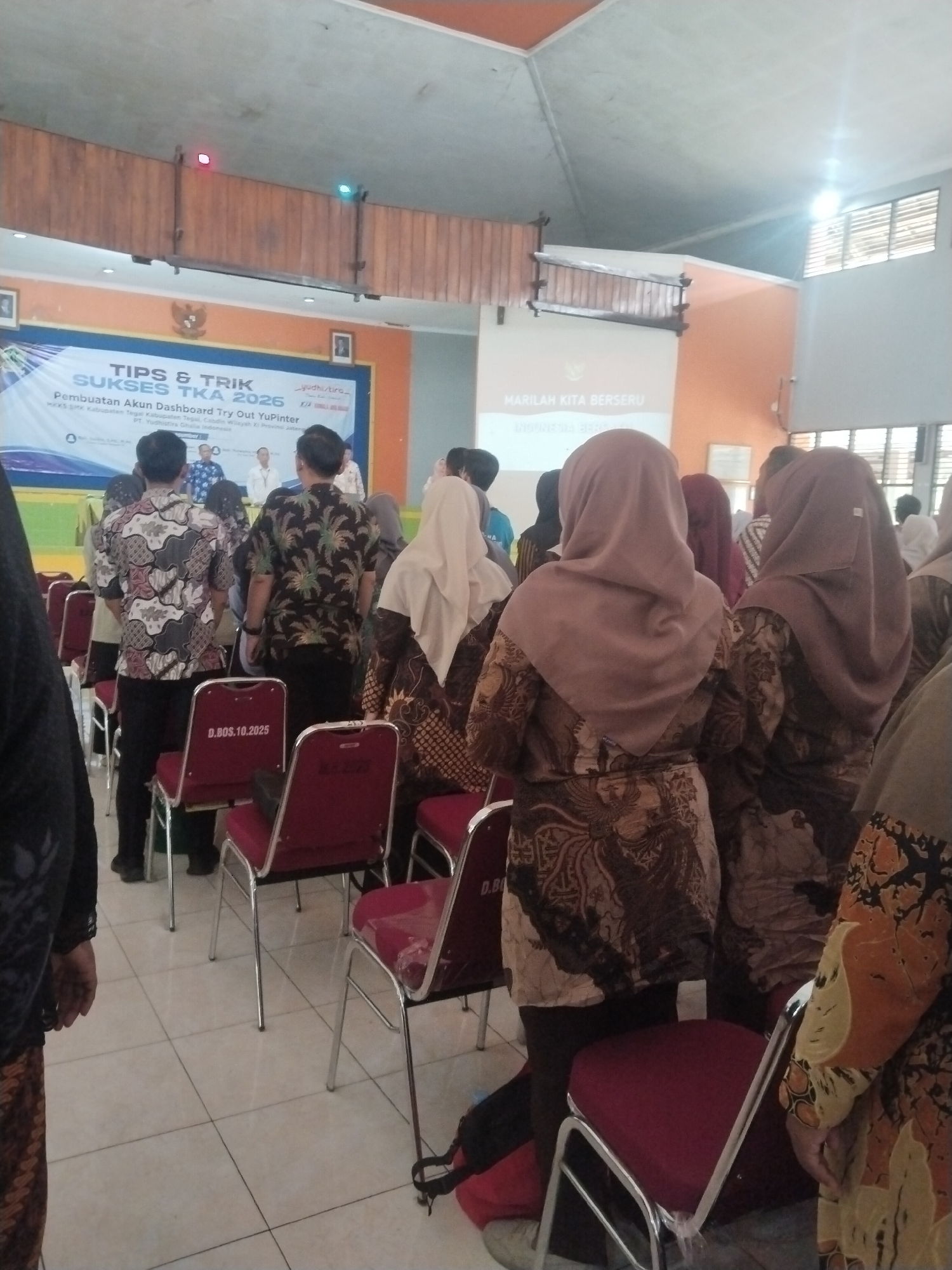 This image depicts an indoor event, likely a seminar or presentation, at the SMA Negeri 1 Pesarean in Pesarean, Indonesia. The room features a stage area with a projector screen displaying "TIPS & TRIK SUKSES TKA 2026" and "PEMBUATAN AKUN DASHBOARD TRY OUT YUPINTER". Several people are gathered, mostly facing the stage. Many attendees are wearing traditional Indonesian batik attire. In the foreground, a row of red chairs with plastic coverings is visible, some with numbers on the backrest like "D.BOS.10.25". The environment suggests a community or educational gathering space.