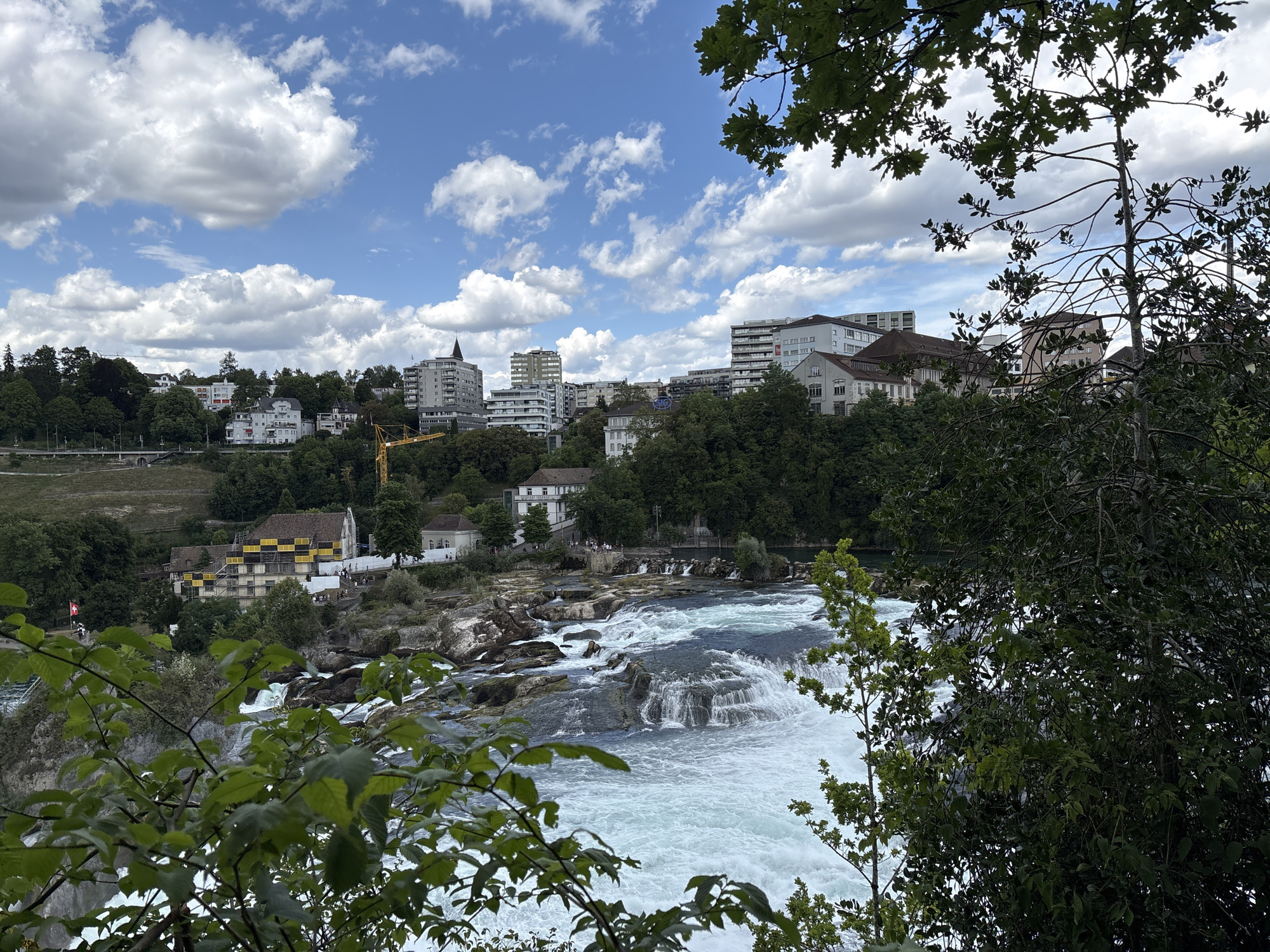 The image depicts a wide, powerful river flowing over a series of rocky ledges, creating a waterfall. The water is a vibrant turquoise blue, churning with white foam. In the background, a town or city is visible, with numerous buildings of varying heights, including residential and taller structures. Trees line the riverbanks and dot the landscape. The sky is blue with scattered white clouds. In the foreground, lush green foliage partially obscures the view of the river and the town. A Swiss flag is visible on a pole near some buildings. A yellow construction crane stands out against the skyline of buildings. The scene appears to be a daytime, natural landscape with urban elements.