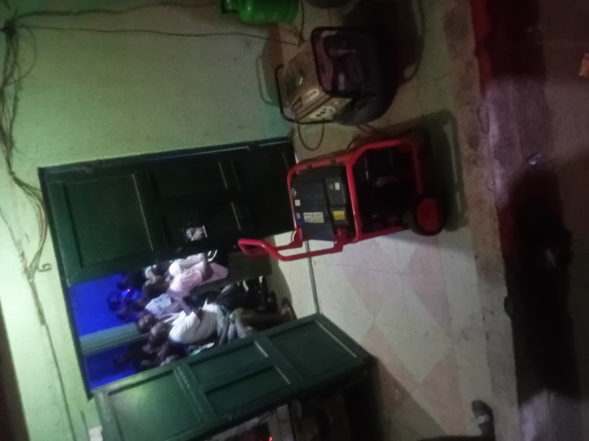 Here is a detailed description of the image:
**Scene:**
The image appears to be taken indoors. There is a door that is open, providing a view into another room. The room visible through the door has a group of people inside, illuminated by what appears to be blue light. To the right of the door is a red generator. Next to the generator is another machine, its color is a darker shade. A green gasoline container is located close to the other two machines. Some wires are hanging from the ceiling on the left side of the door. A green object with a box shape is near the right part of the door frame. The environment looks dimly lit.
**Subjects/Objects:**
* People: There is a group of people visible through the doorway. Their specific actions are not discernible.
* Door: A partially open green door.
* Generator: A red generator is positioned near the door.
* Gasoline container: A green gasoline container located close to the other two machines.
* Another machine: Near the red generator is another machine with a darker shade.
**Location:**
* City: Unknown
* State/Province: Unknown
**Environment:**
* The location is indoors and dimly lit.
* The condition and setting suggest a rudimentary or possibly temporary setup.