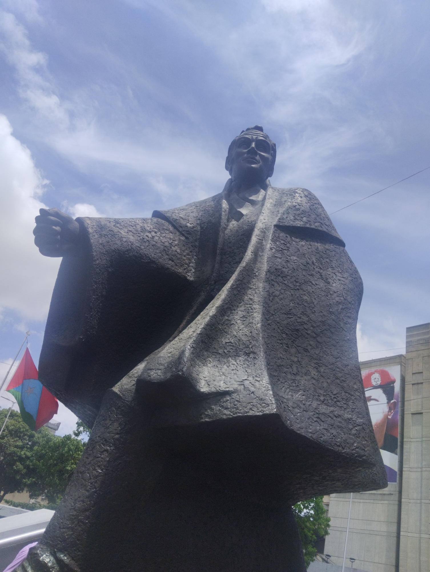 The image shows a bronze statue of Venezuelan political leader Rómulo Betancourt, located in Caracas, Venezuela. Betancourt is depicted wearing a long robe or cloak, with his right hand clenched in a fist. In the background, there is a building with a large poster of Hugo Chávez, also a former Venezuelan president. A Venezuelan flag is visible to the left of the statue. The sky is blue with scattered clouds. The shot is taken from a low angle, emphasizing the statue's grandeur.