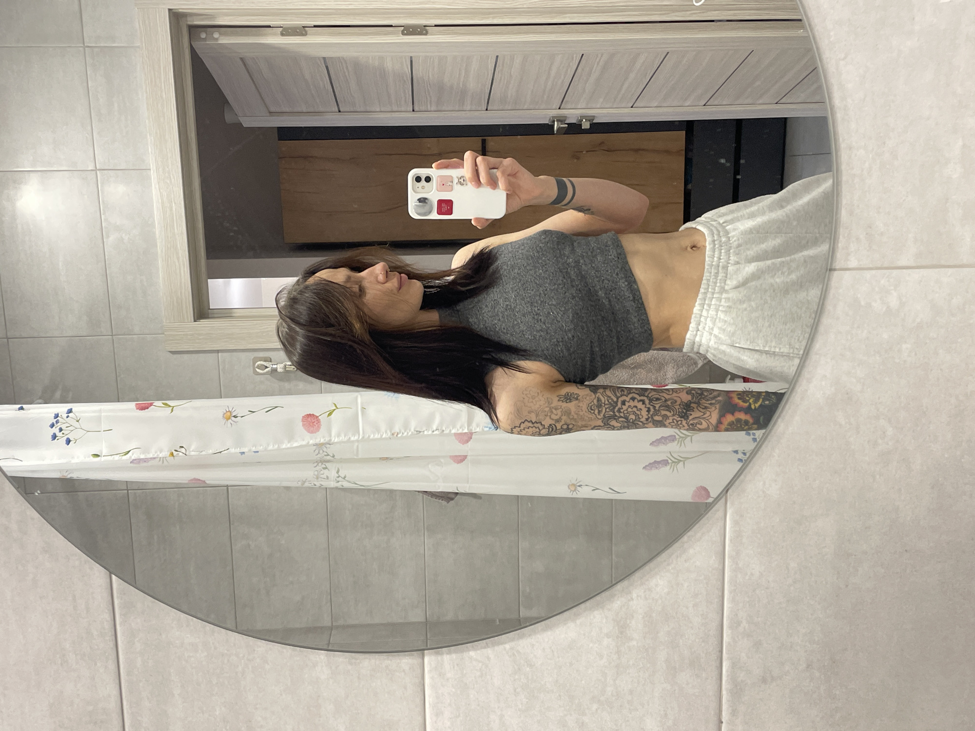 A young woman is captured in a mirror selfie inside a bathroom. She stands, reflected in an oval-shaped mirror, holding a white smartphone with multiple colorful attachments/stickers to take the photo. Her dark hair is long and falls over her shoulders. She wears a dark gray crop top, revealing her midriff, and light gray sweatpants. Notable features include a nose piercing and extensive black-ink tattoos covering her left arm, along with thick black armbands on her right arm.
The bathroom features light gray tiled walls. Behind the woman, the reflection shows a light wooden framed cabinet or vanity with light gray paneling above. A shower curtain with a delicate floral pattern, featuring blue, pink, and purple blossoms, is visible to the left and below her in the mirror's reflection. The overall setting is clean and modern. The image provides no specific time of day or emotional cues beyond the act of taking the selfie. No text is visible within the image. This scene takes place in Izhevsk, Russia.