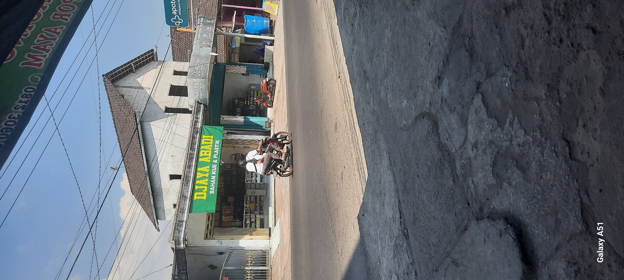 The image captures a street scene in Selopuro, Indonesia, under a clear, sunny sky. In the foreground, a damaged asphalt road with visible cracks and patches is present. A motorbike carrying two individuals, a man and a woman, travels on the road from left to right. The man is driving, and the woman is seated behind him. They are both wearing casual clothing.
To the left of the road, a building houses two storefronts. The left storefront, branded "DJAYA ABADI BAHAN KUE & PLASTIK," displays a green banner and has its interior filled with shelves stocked with various items. The right storefront, identified as an "apotek selopuro farma," also features shelving and merchandise visible through its open entrance. A red scooter is parked in front of the apotek. The building's facade is light-colored with visible windows.
In the background, another building with a tiled roof is visible. Overhead, numerous power lines crisscross the sky. A sign with text including "MAYA RO" is partially visible at the top of the image. A blue tarp covers an object on the right side of the scene. A sign for "SERVIS" is also present on the right. The overall environment suggests a commercial area in a town.