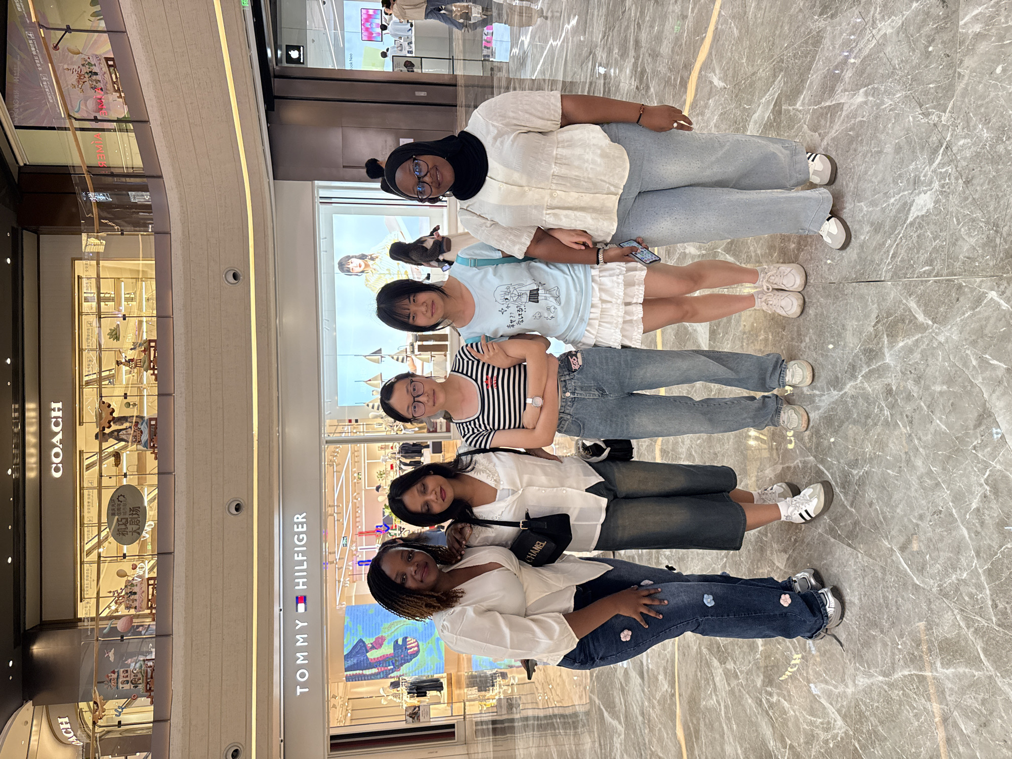 Five women are posing for a photo in a brightly lit, modern indoor shopping mall. The women are standing on a polished marble floor. In the background, there are storefronts, including a Tommy Hilfiger store. Above the storefronts, there is a second level with more storefronts and illuminated displays. The overall environment suggests a high-end retail setting in a contemporary urban area.