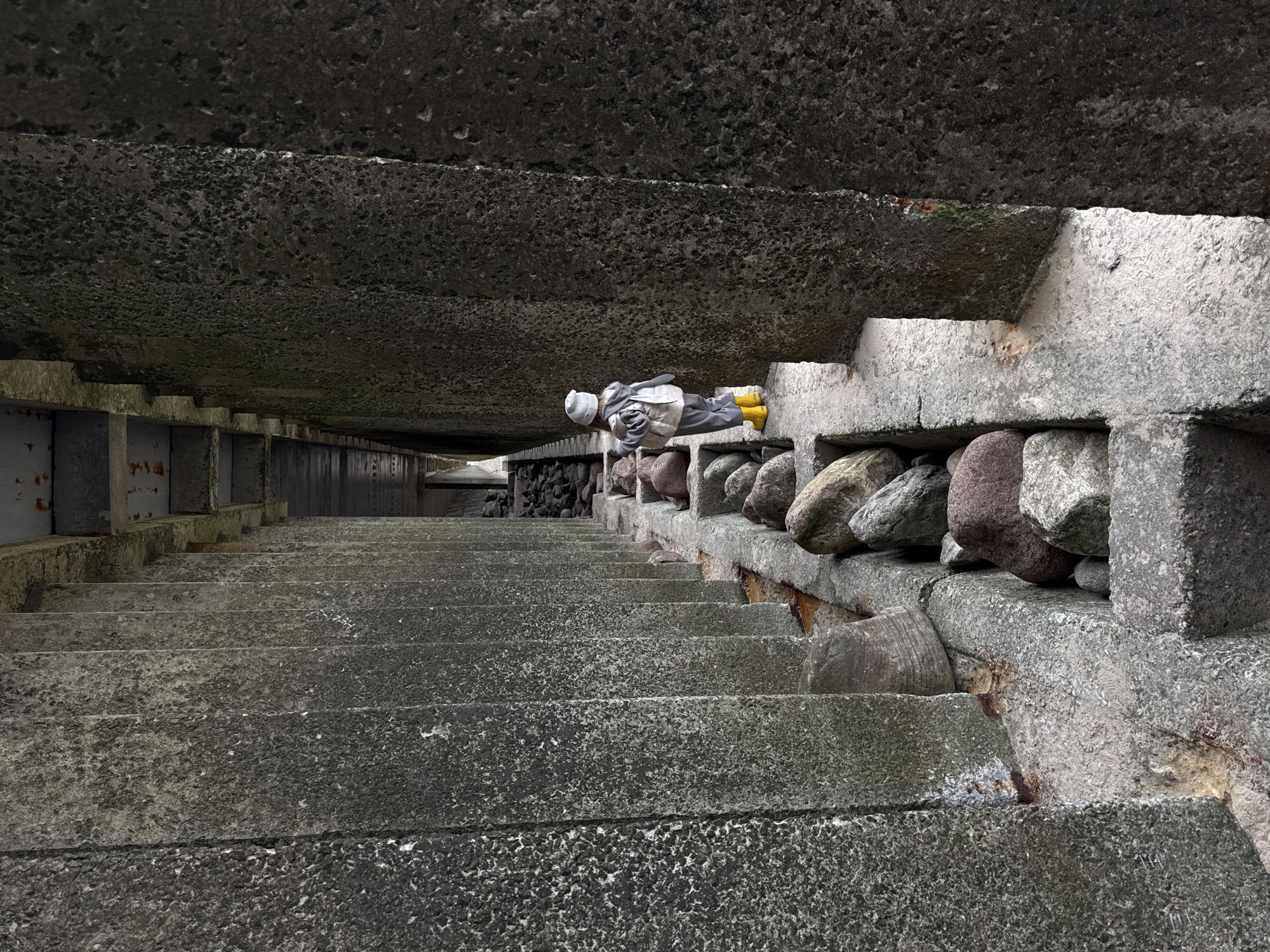 The image depicts a long, concrete staircase in an urban setting, seemingly in the city of Zelenogradsk, Russia. The staircase ascends into shadow with a gray concrete ceiling overhead. The right side of the staircase has a rough, stone wall built up along the edge, with several large, rounded rocks forming a barrier. A person lies on the edge of the stairs near the top of the visible section. They are wearing a light-colored hat, gray jacket, and yellow boots. The person is lying on their side facing into the stairs. The left side of the staircase is a concrete structure with rectangular openings along the side, resembling a corridor or walkway. At the top of the stairs, a wooden fence or gate can be seen. The entire scene is dominated by shades of gray, suggesting overcast or dim lighting. The texture of the concrete and stone is rough and weathered.