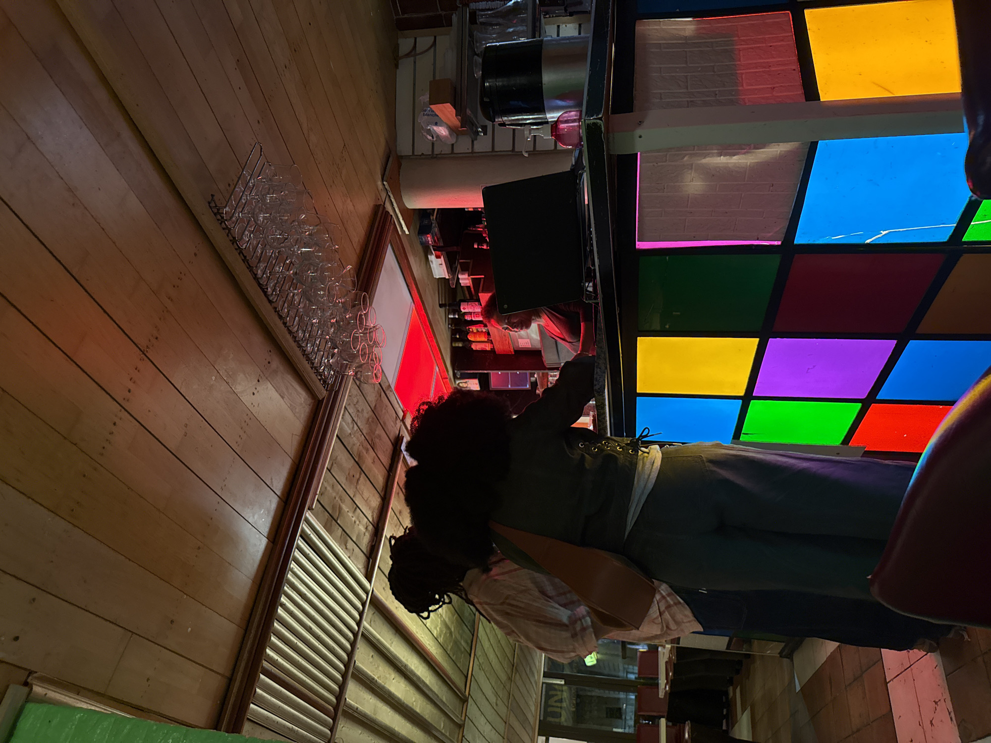 A dimly lit interior shot from a bar or restaurant, potentially in Paris, France, given the ambiance and possible presence of French-language signs. The perspective is unusual, seemingly taken from near the ceiling or high on a wall, rotated 90 degrees counterclockwise. The camera appears to be looking down and slightly to the right.
A wooden wall and ceiling are visible on the left, featuring horizontal planks and a wine glass rack mounted high. A woman with dark, curly hair is standing behind a bar. She's wearing an olive-green jacket, a light-colored plaid shirt, and dark pants. She appears to be interacting with the bar area. The bar itself has a variety of bottles and equipment on display, possibly for preparing drinks.
A colorful stained-glass-style window panel, composed of multiple squares in red, blue, green, yellow, and purple, is visible to the right of the bar. The overall environment suggests an older, somewhat eccentric establishment with a unique character. There is also another set of patterned panels below.