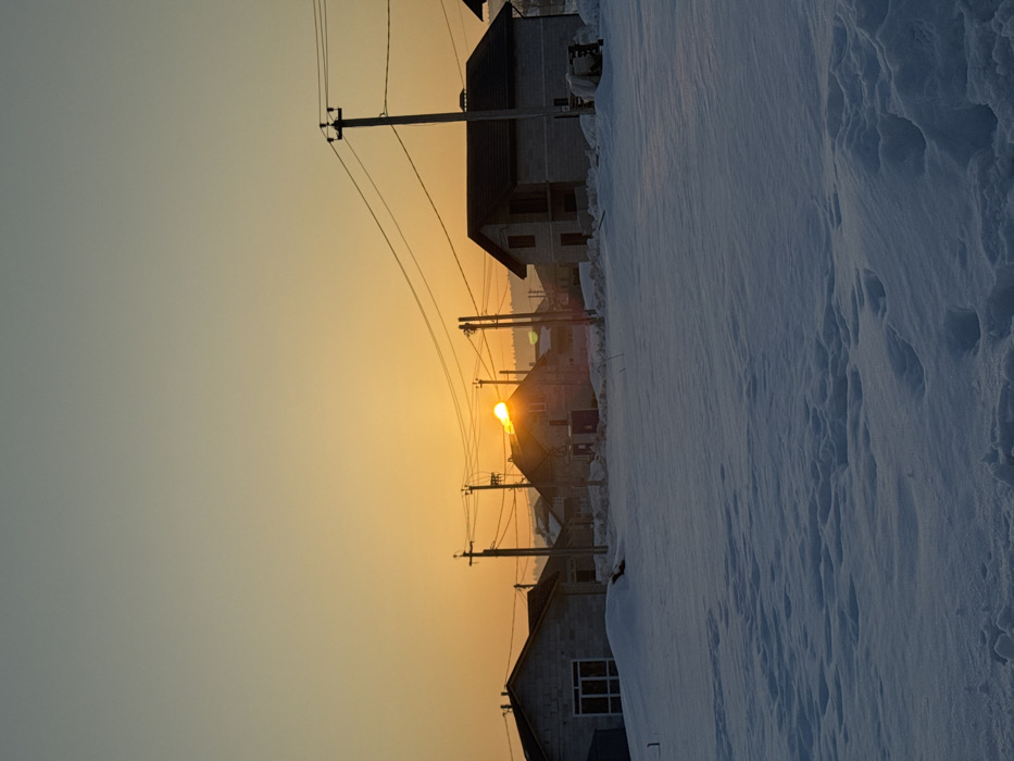 The scene depicts a snow-covered landscape in Gornyi (Dvurechenskoe S/P), Russia, during a sunrise or sunset. The foreground consists of a field of undisturbed snow, showing variations in depth and texture. In the midground, a row of residential buildings is visible. The prominent structure on the left is a single-story building under construction, featuring exposed light grey blockwork walls and a large, dark-framed window on its visible side. Further right, a two-story house with similar grey blockwork walls and a dark sloped roof is partially obscured. The sun, a bright orange orb, is low on the horizon, partially hidden behind a central house, casting a strong glare. Numerous wooden utility poles with multiple overhead power lines traverse the midground horizontally across the image. The background features more distant houses and a faint outline of hills or mountains. The sky transitions from a deep orange at the horizon, where the sun is located, to a pale blue-grey at the top of the frame. No individuals or animals are present in the frame.