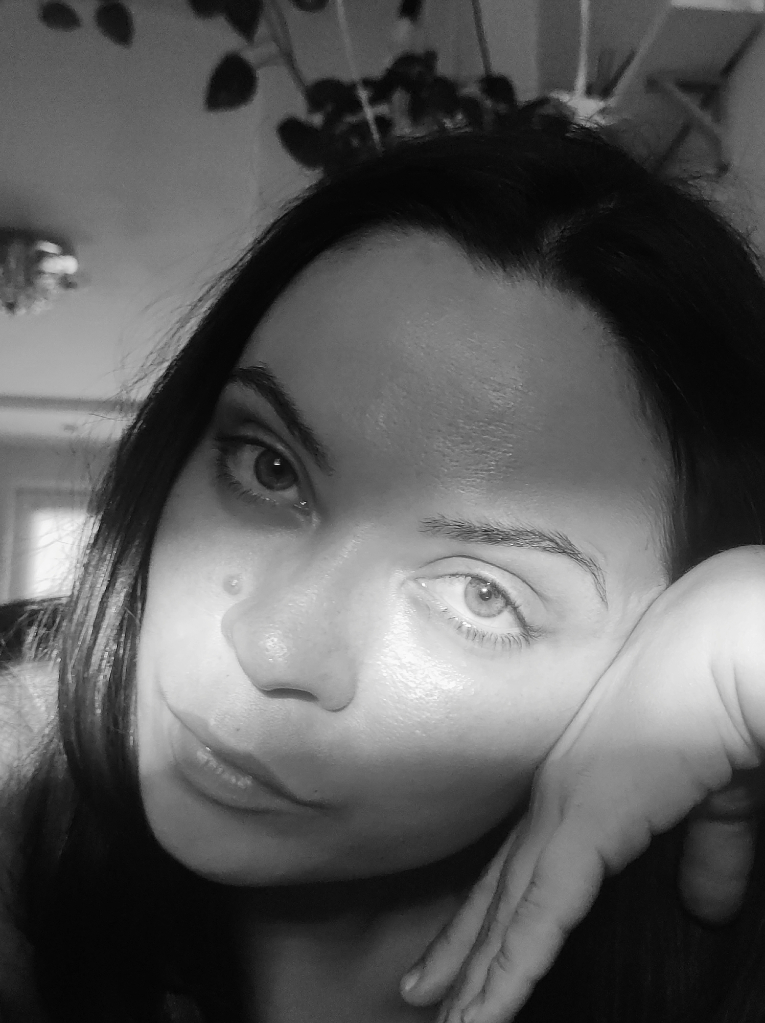 This is a black and white, close-up photograph of a woman's face, with her hand resting on her cheek. She has dark hair and her left eye is looking towards the camera, while her right eye is partially obscured by her hand. There is a shadow across her forehead and cheek, suggesting a light source from the side. A small, round blemish is visible on her cheek. Her lips are slightly parted. The background is blurred but appears to be an indoor setting with some foliage and furniture visible, possibly a home. The overall mood of the image is contemplative or introspective. There is no visible text. The location context is Chaikovsky, Russia.