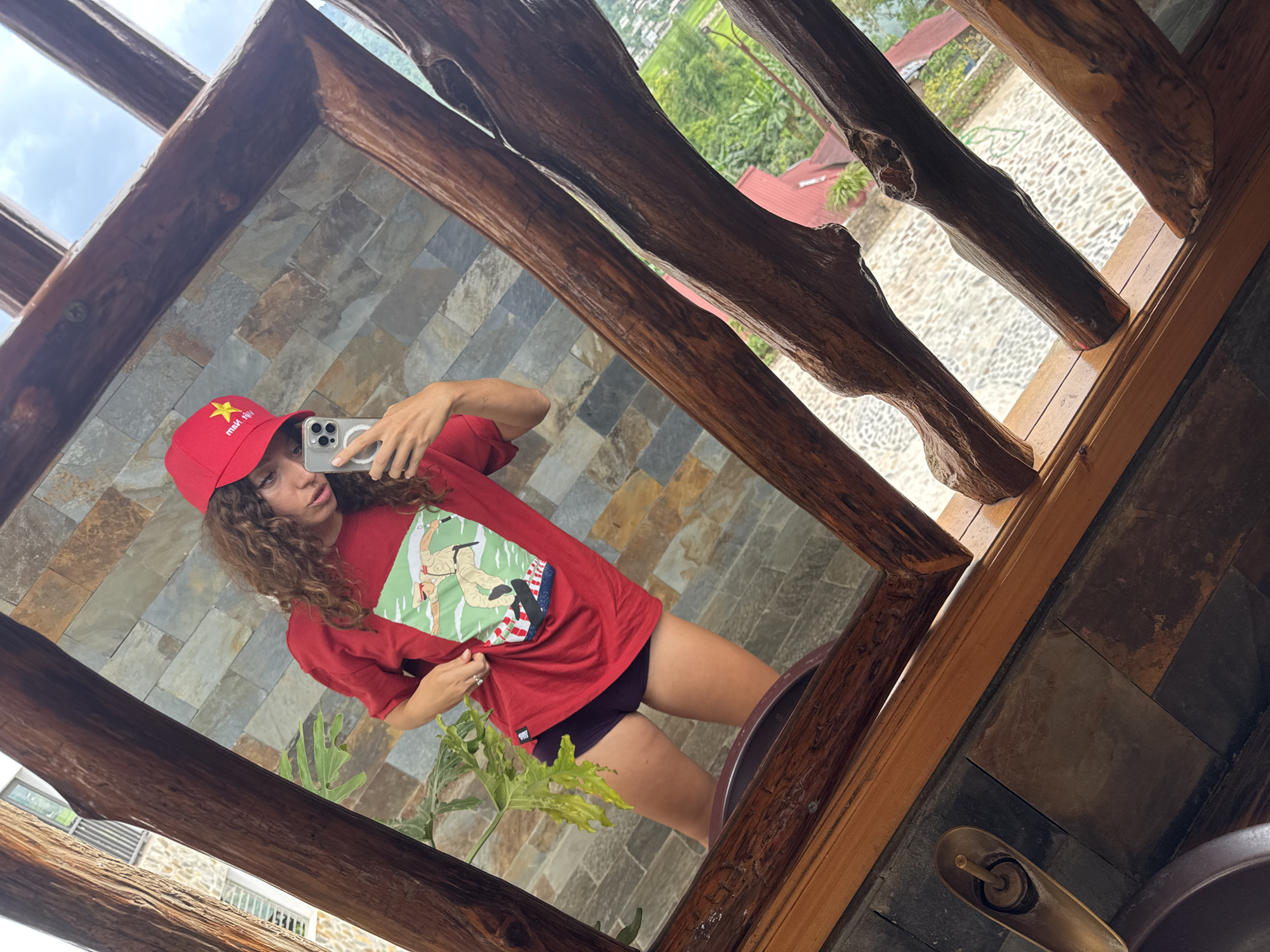 A young woman is taking a mirror selfie in what appears to be a bathroom in Tú Lệ, Vietnam. She is wearing a red baseball cap with a yellow star, a red T-shirt with a graphic, and dark-colored shorts. The mirror has a unique wooden frame made from tree branches. In the reflection, stone walls are visible, and a plant is near the sink. Outside the window, a view of the landscape with buildings and greenery typical of the Tú Lệ area can be seen. The sink in the bathroom has a metal faucet and is built into a surface made of stone tiles.