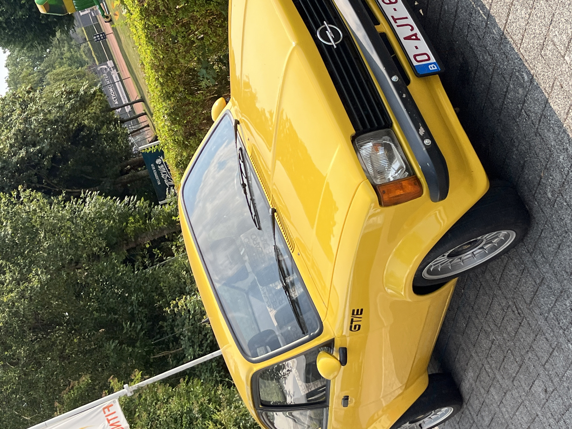 A bright yellow Opel GT/E is parked on a brick-patterned surface. The car features a black grille with the Opel logo, distinctive headlights, and the GT/E badging on the side. The license plate reads "B O-AJT-6". In the background, there is a lush green hedge and trees, and beyond that, a tennis court with fencing and advertising banners, one of which reads "ACTIONCENTER". A white flag with red lettering is visible to the left of the car. The overall scene suggests an outdoor event or gathering in Saint Vith, Belgium.