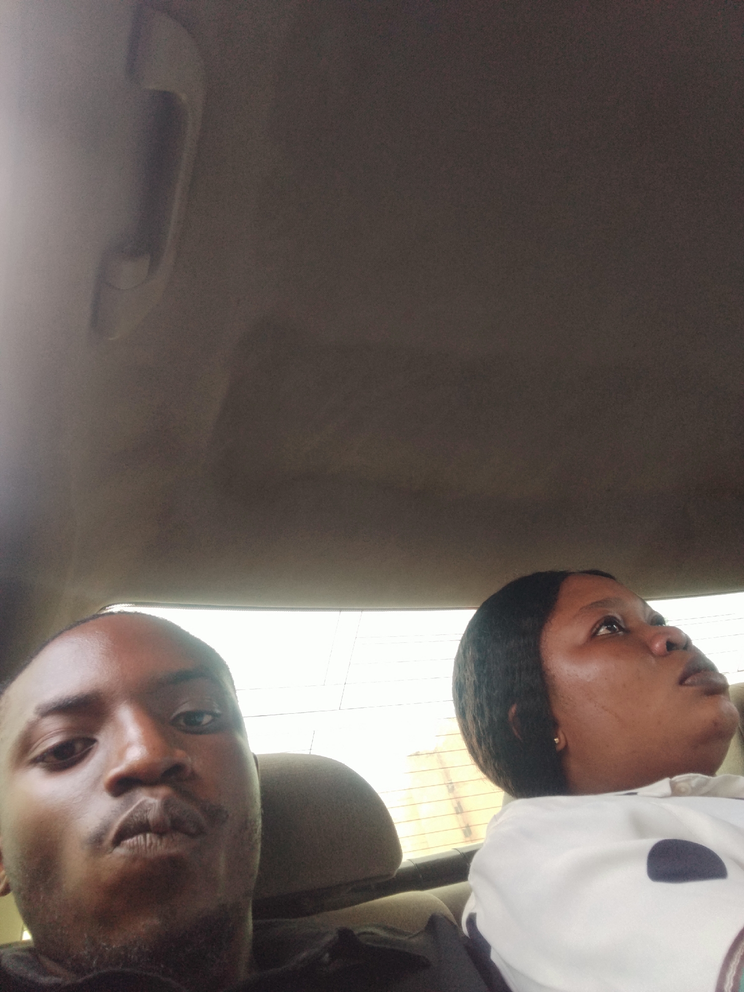The image shows a close-up of two people in a car, presumably in Abuja, Nigeria. On the left, a man is looking directly at the camera, his face filling the foreground. He has short hair and appears to be wearing dark clothing. To the right, a woman is looking upwards, her head tilted. She has dark hair and is wearing a white shirt with dark polka dots. The background is the interior of a car, with the headliner and a grab handle visible. The window behind the woman shows a blurred outdoor scene, suggesting daylight. The overall mood seems casual, possibly a selfie taken during a car ride.
