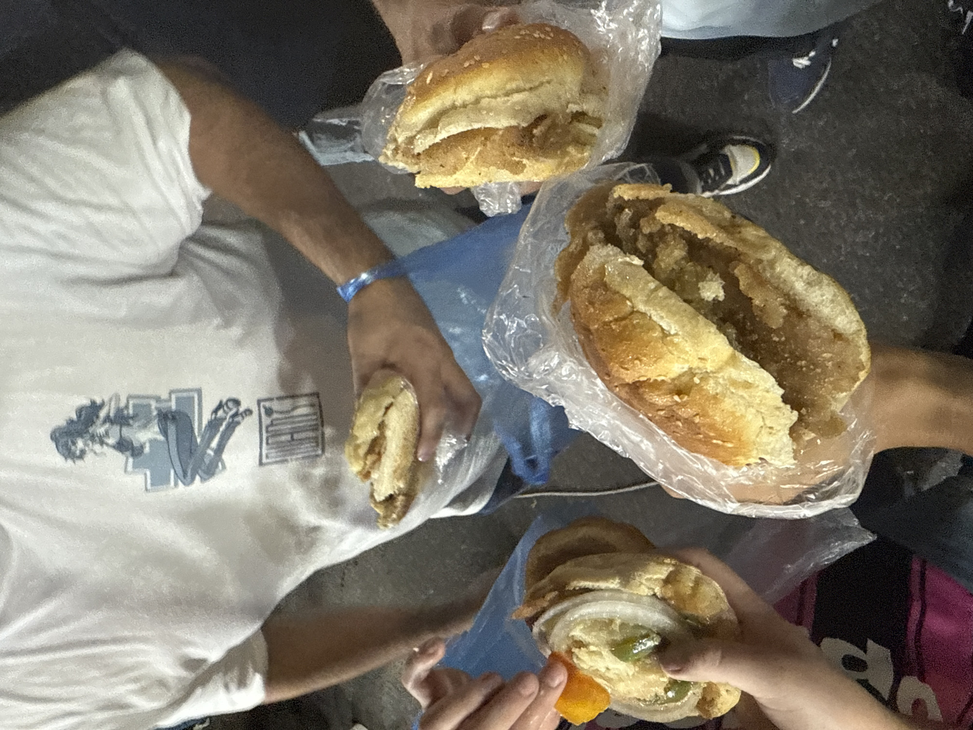 The image shows four people holding individually wrapped sandwiches or "cemitas" in what appears to be an outdoor setting. The cemitas are in clear plastic bags, with the bread of each sandwich visible. One person is wearing a white t-shirt with a graphic print on it, and another has a blue wristband. The background is dark and grainy, possibly indicating an asphalt surface. The image is taken from an overhead perspective. Due to the visible "cemitas," it is possible that the photo was taken in Puebla, Mexico, a city known for the dish.