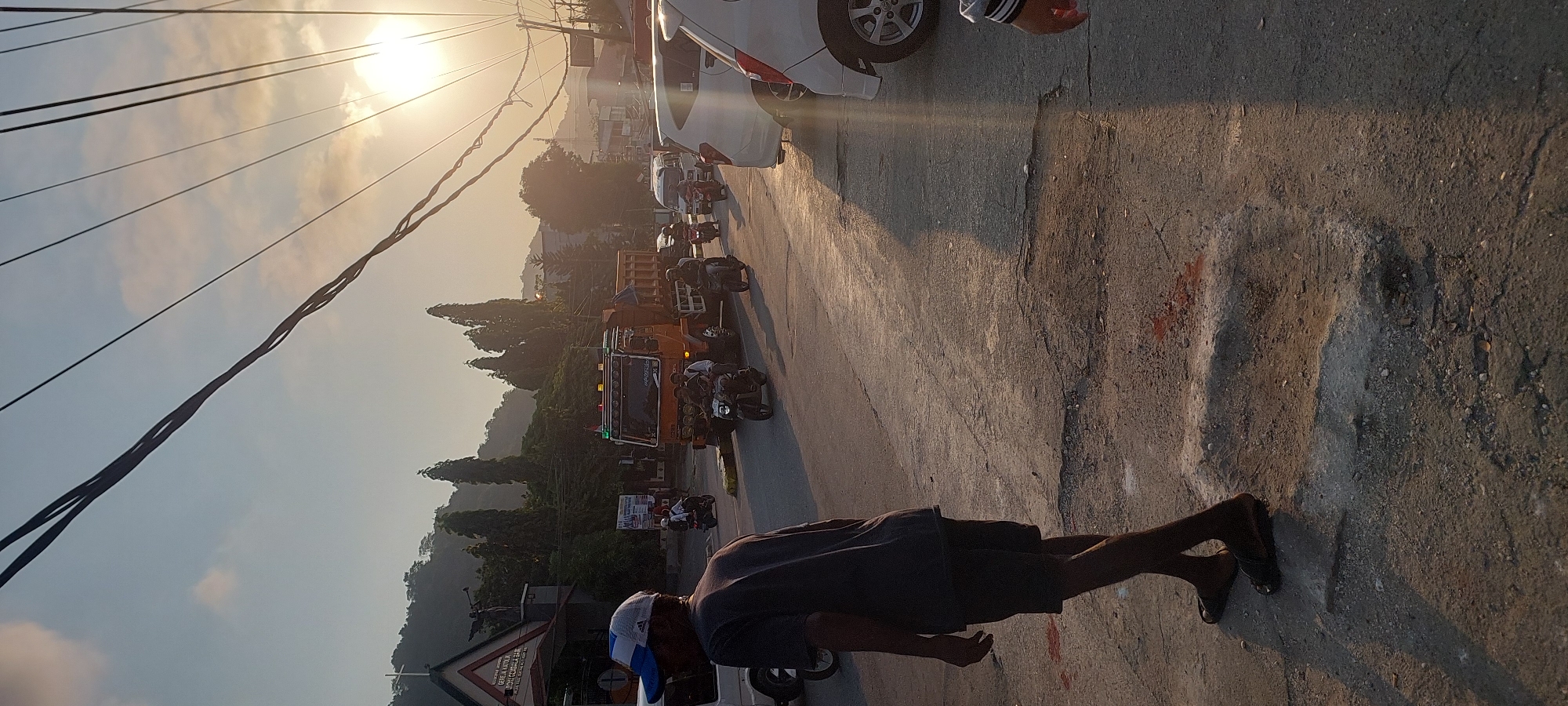 In Jayapura, Indonesia, at approximately 6:00 PM, a street scene unfolds under the warm glow of the setting sun. The image captures a man in a grey t-shirt and shorts walking away from the camera on a cracked asphalt road. He wears a blue and white baseball cap and sandals. In the background, a busy intersection is visible. A large orange truck with its lights illuminated is positioned centrally, followed by several motorcycles and a white van. The road is lined with trees and buildings, with a church steeple prominently visible to the left. Power lines crisscross the sky, and the sun's rays create strong shadows across the pavement. The overall impression is of a typical evening in a bustling Indonesian city.