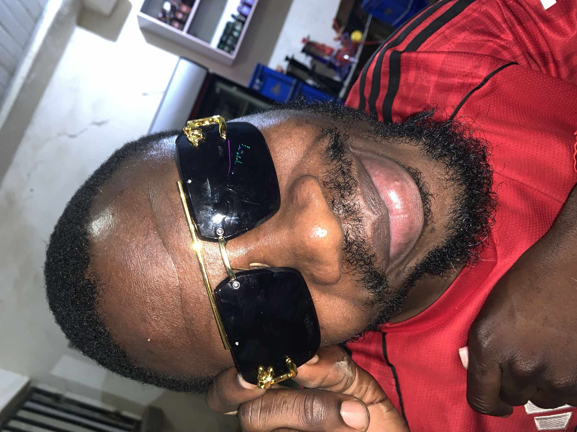 A close-up, waist-up, forward-facing shot shows a Black man from the chest up. He is wearing a red shirt with black stripes on the shoulders. He is also wearing gold-rimmed sunglasses with black lenses. He is holding the side of his sunglasses with his left hand. His mouth is open, and he has a beard. The background is out of focus but appears to be an indoor setting, possibly a shop or market stall. There are shelves with items on them in the background. The lighting is bright and appears to be artificial. The man's skin tone is dark.