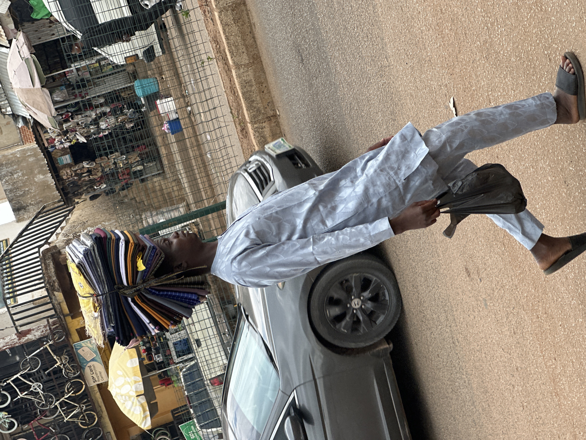 The image shows a man walking along a street next to a grey car. The man is carrying a stack of colorful fabrics on his head and holding a dark bag in his hand. He is wearing a light-colored, long-sleeved shirt and pants, and slides. The location appears to be an urban street in Bukuru, Nigeria, with shops and other bicycles visible in the background.