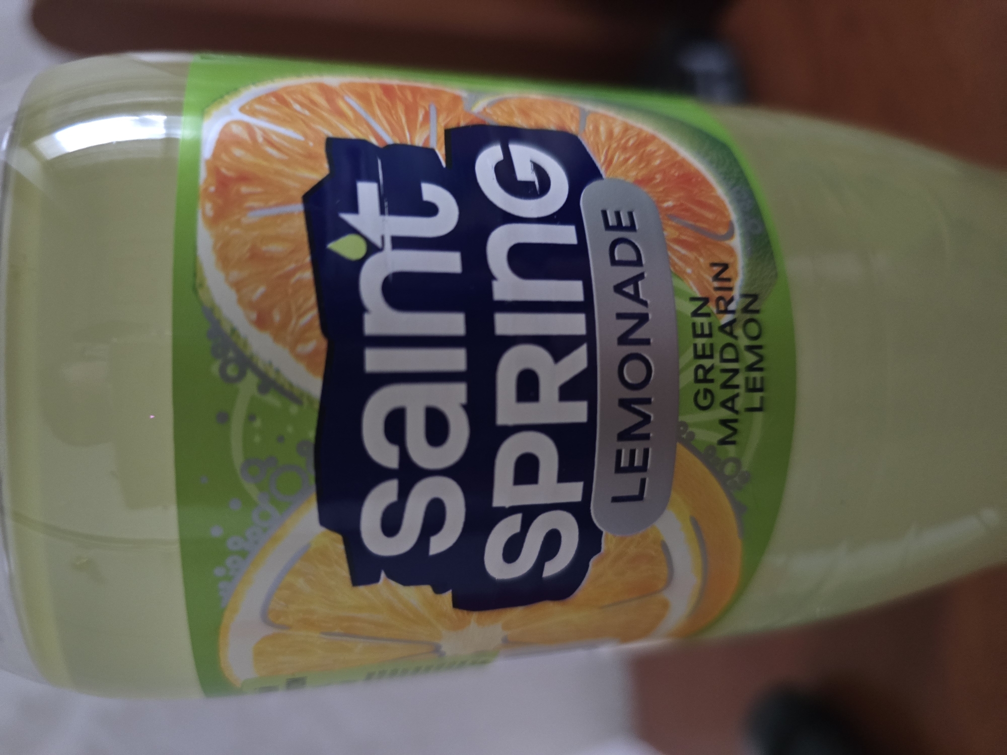 This media file shows a close-up, angled shot of a bottle of Saint Spring Lemonade. The bottle has a light green liquid inside and is adorned with a label featuring citrus fruits, specifically orange slices. The label has a blue background with white text that reads "SAINT SPRING" in large, bold letters. Below this, "LEMONADE" is written in smaller, capitalized text. To the right of "LEMONADE," the text "GREEN MANDARIN LEMON" is visible in green lettering. The overall aesthetic is bright and refreshing, suggesting a summer beverage. The background is blurred, with hints of a brown surface, possibly a table or countertop. The lighting suggests an indoor setting, likely during the day. There are no people or animals visible in the frame.