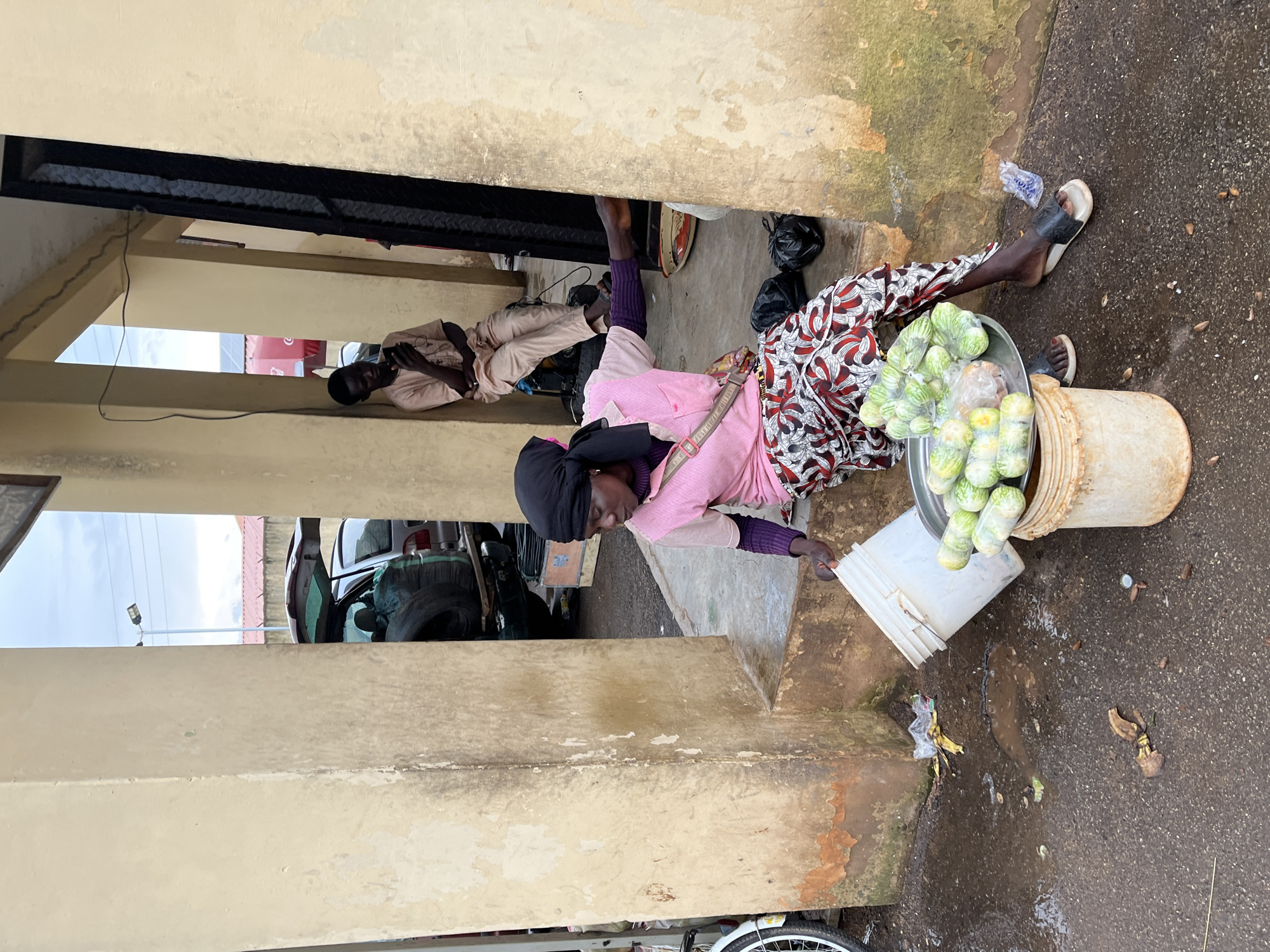 In Bukuru, Nigeria, a woman sits on a concrete step in front of a building with beige, weathered walls and columns. She wears a black headscarf, a pink long-sleeved shirt, and a patterned skirt. In front of her, a metal tray filled with wrapped produce, likely vegetables, rests on top of a white bucket. Another white bucket, tipped over, lies beside it. To her right, two black plastic bags are on the ground. Behind her, a man in a light-colored robe sits against a wall, looking at his phone. Further in the background, a car with its trunk open is visible, with tires and other items inside. The ground is wet and appears to be a mixture of concrete and dirt, with scattered debris. The scene suggests an outdoor market or roadside selling area.