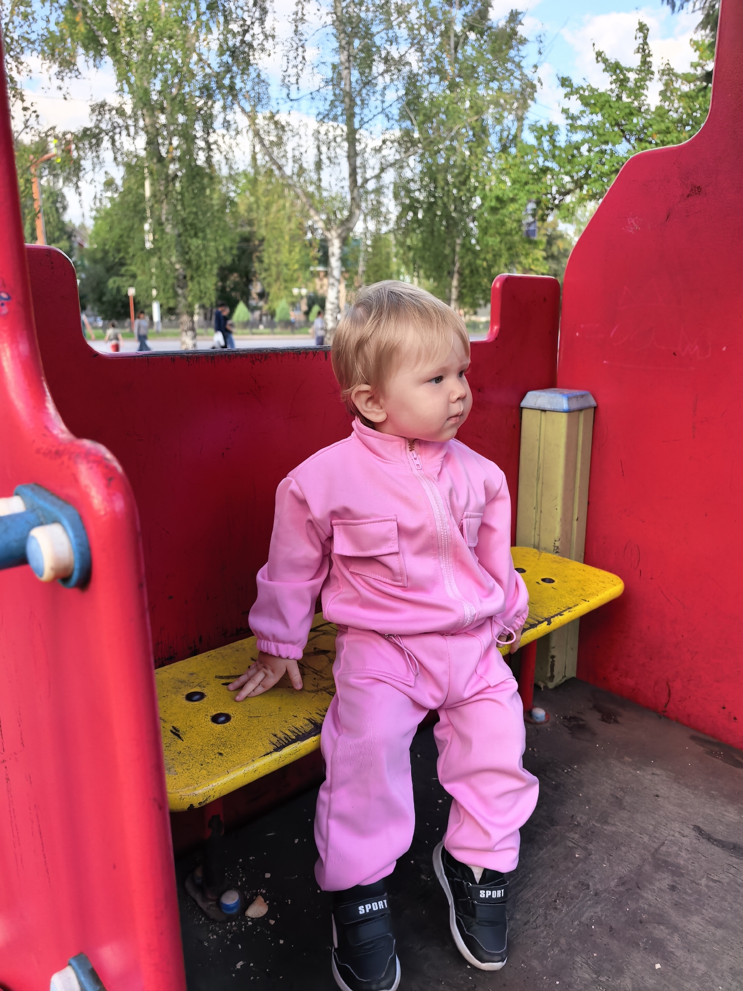 A young child, with blonde hair and fair skin, sits on a yellow bench within a red structure, possibly part of a playground. The child is wearing a pink tracksuit and black athletic shoes with white "SPORT" lettering. The child is looking to the right of the frame. In the background, trees with green foliage are visible under a blue sky with scattered clouds. Several blurred figures of people can be seen in the distance. The ground appears to be made of dark material, with some debris on it. The setting suggests an outdoor public space, like a park or playground, potentially in Sterlitamak, Russia given the context.