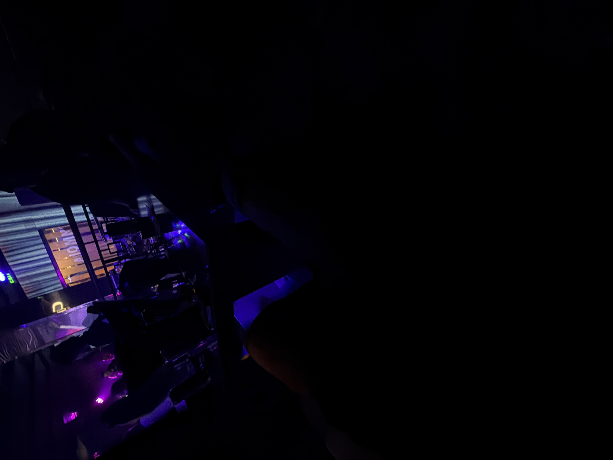 The image shows the interior of a dark room, possibly a bar or club, likely in Sofia, Bulgaria due to the neon sign featuring the word "ATOMIX," which is the name of a Sofia bar. The room appears to have tables and chairs, and there is stage lighting present, including purple and blue hues, suggesting a live performance space. The scene is dimly lit, and most of the right side of the image is obscured by darkness.