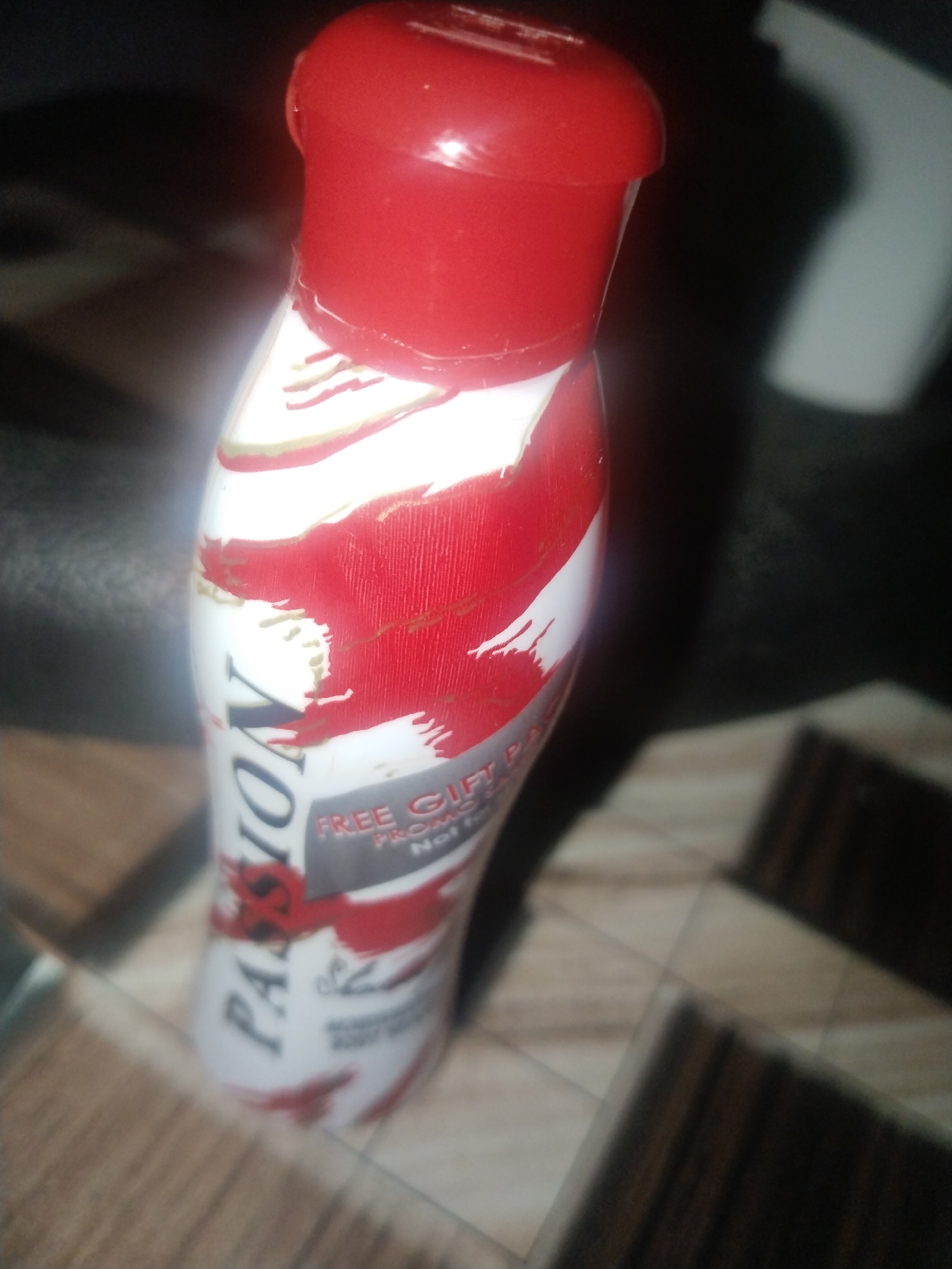 The image depicts a bottle of "Passion" body wash. The bottle is white with red accents and a red cap. The text on the bottle includes "FREE GIFT PACK Promotion." The location is unclear, only that the bottle sits on a surface composed of brown and beige tiles.