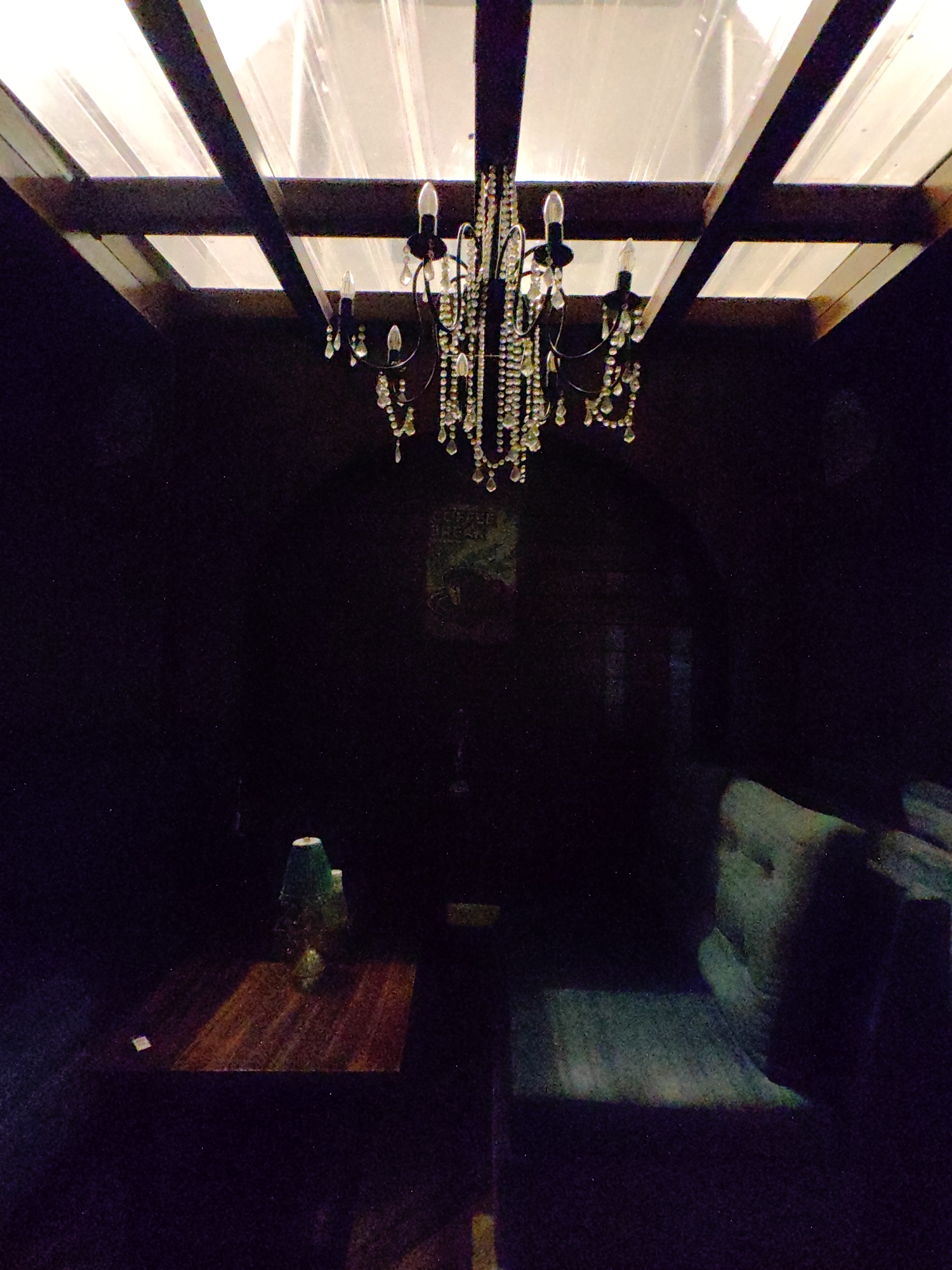 A chandelier with multiple lights and hanging crystals is visible above a wooden table with a lamp on it. In the background, a dark wall features a poster. To the right, a seating area with cushions is depicted. The scene is dimly lit, suggesting an indoor setting, possibly in Barheta, India, though this cannot be definitively confirmed from the image alone.