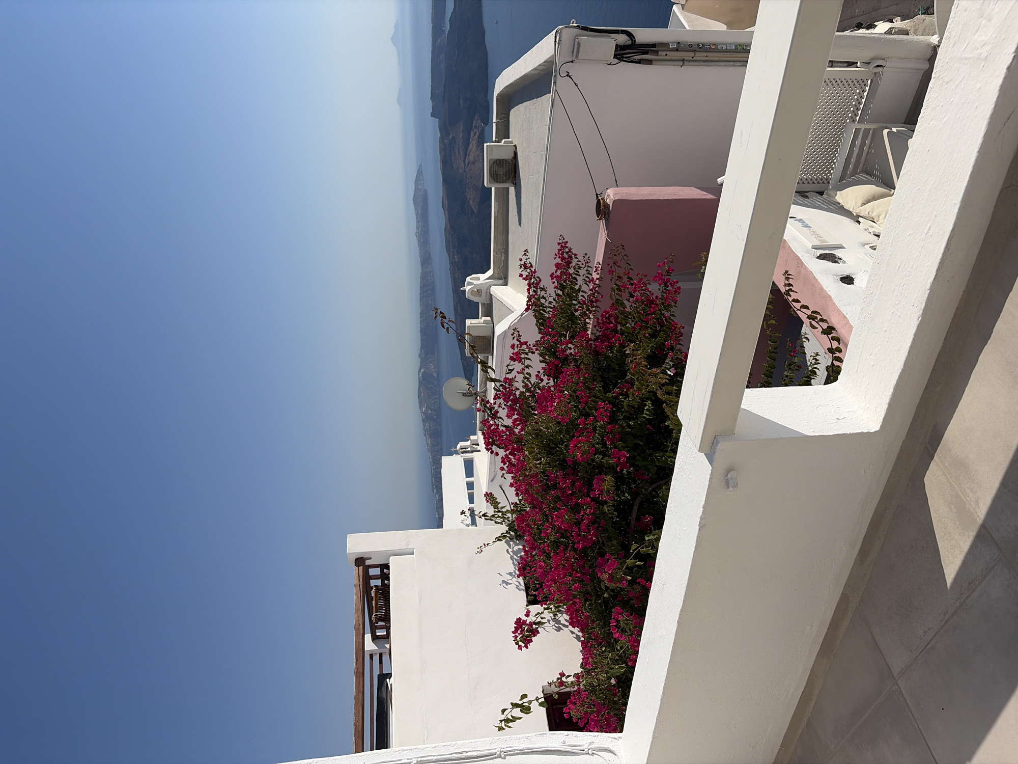The image displays a view of a white-railed balcony overlooking a seascape in Thira Municipal Unit, Greece. The foreground features a tiled floor and a white railing. Beyond the railing, a vibrant pink bougainvillea bush with dark green leaves is prominently situated against a white building. In the middle ground, more white buildings with flat roofs are visible, some equipped with air conditioning units. A satellite dish is also present on one of the rooftops. The background consists of a clear blue sky merging into a hazy horizon, with several islands in the distance across a blue body of water. The overall scene depicts a bright, sunny day with architectural elements typical of a Greek island setting.