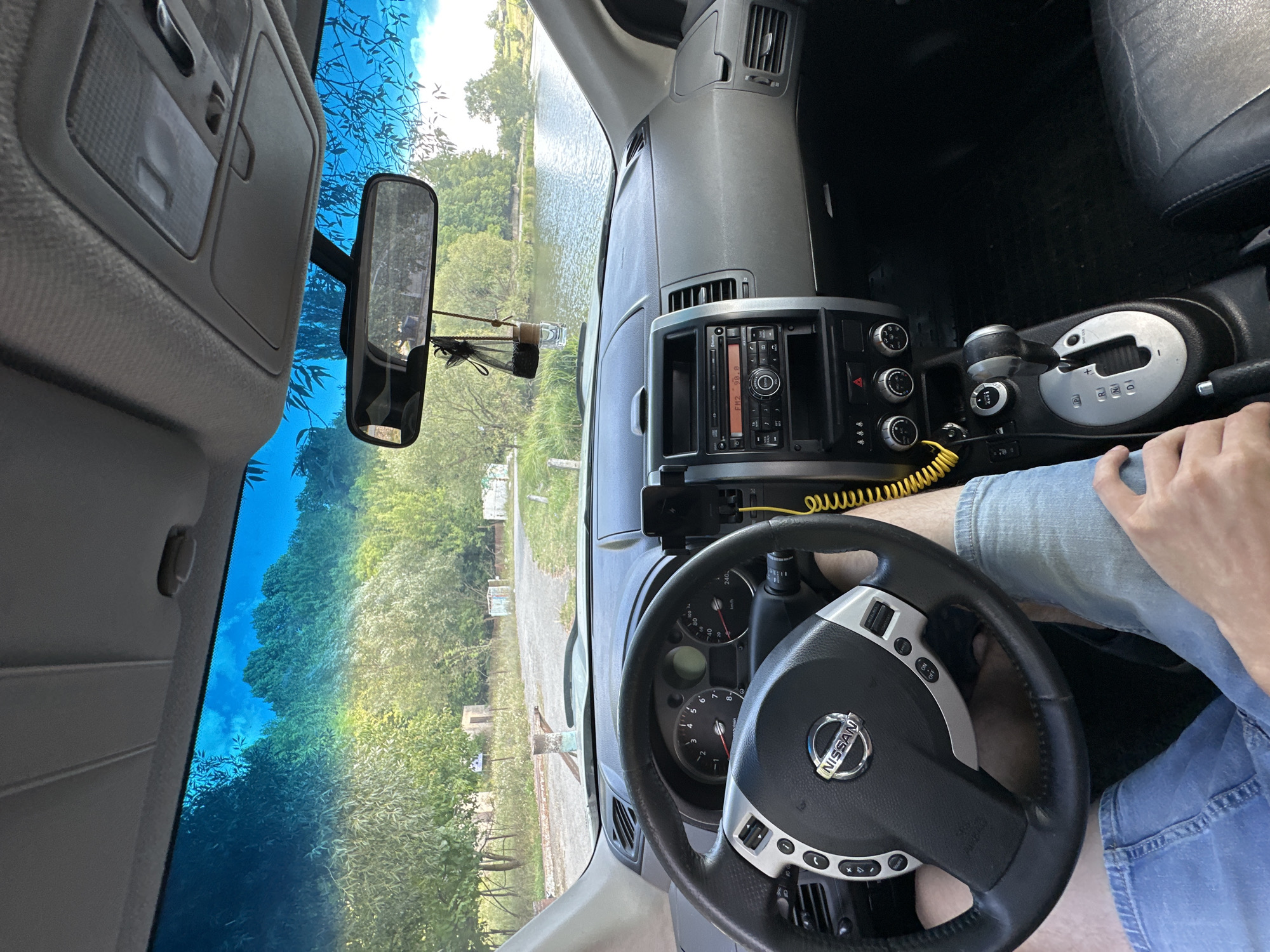 The image shows the interior of a Nissan X-Trail, viewed from the driver's perspective. A person's legs are visible, wearing blue denim jeans. The driver's seat is occupied, and the steering wheel, dashboard, and center console are in focus. Outside the vehicle, a body of water, likely a lake or pond, is visible, surrounded by trees and vegetation. The sky is bright blue with some clouds. The scene appears to be in a rural or semi-rural setting. The location is identified as Makiv, Ukraine.