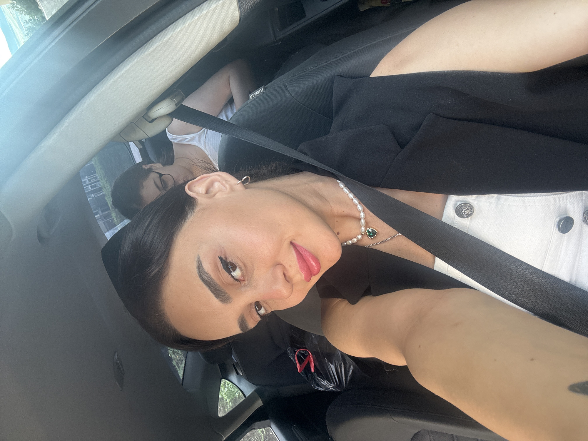 A selfie taken from the front passenger seat of a vehicle shows a woman with dark hair, applying lipstick and looking directly at the camera. She wears a white top and a black blazer, with a pearl necklace and a pendant. Behind her, in the rear seat, another woman with glasses is visible. The vehicle's interior, including the seats and seatbelt, is in focus. Outside the car, trees and buildings can be seen through the windows, suggesting an urban or suburban environment. The scene is consistent with a drive through a city, potentially Tbilisi, Georgia, given the architecture and vegetation.
