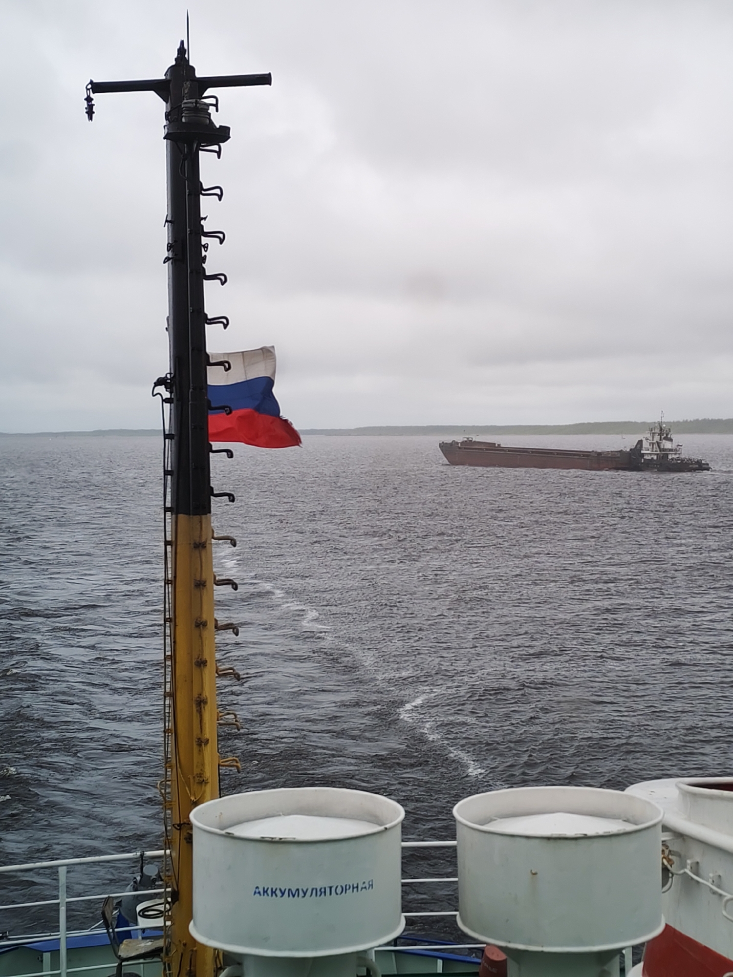 The image shows a view from a ship or boat. In the foreground are two cylindrical containers with the word "АККУМУЛЯТОРНАЯ" written on them. A metal pole with a ladder and a Russian flag attached to it is located to the left of the containers. In the background, there is a body of water with a ship or barge moving across it. The sky is overcast and grey. The location is likely near the city of Gorushka, Russia, given the Russian flag and Cyrillic text.