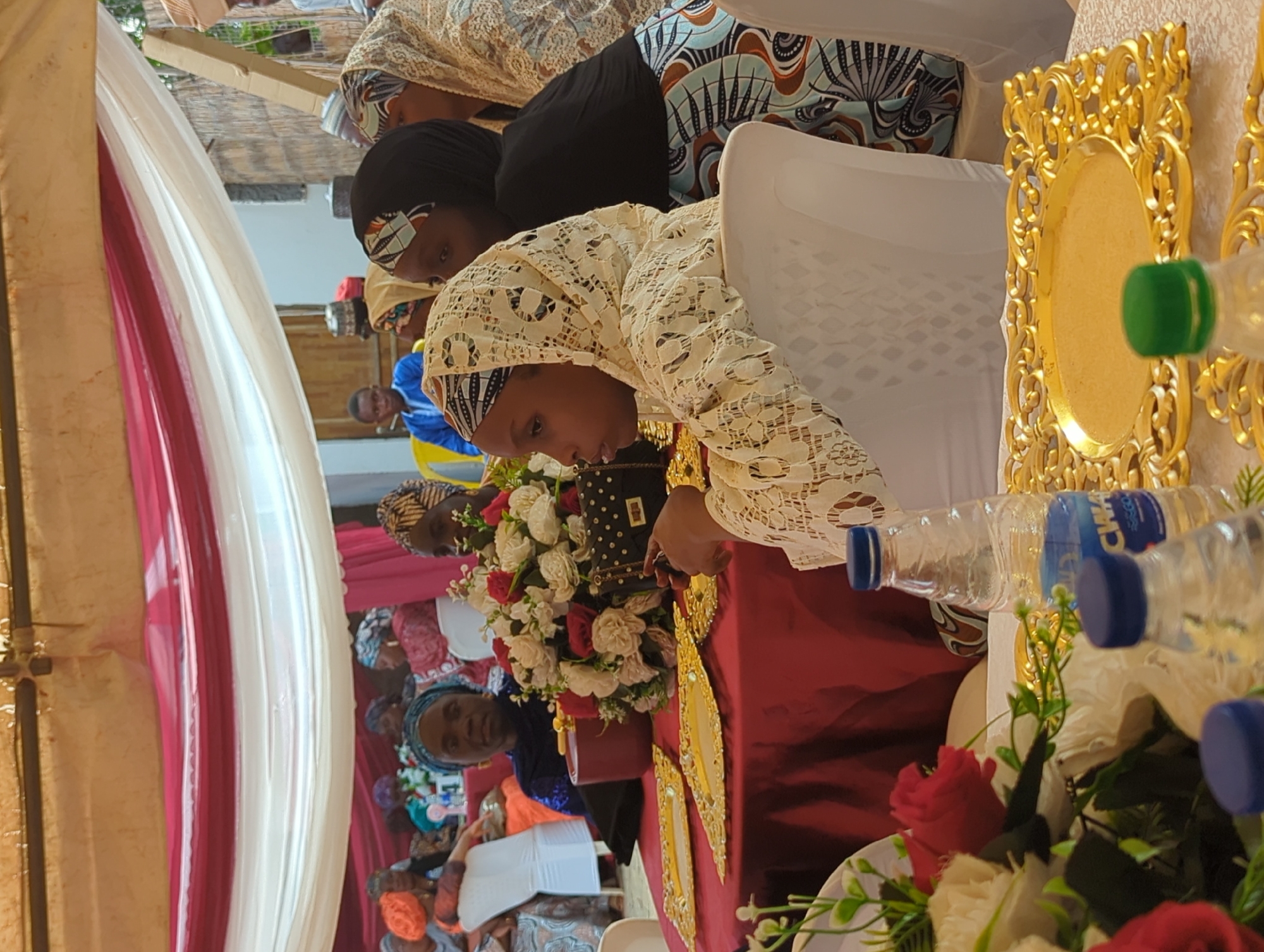 This image captures a formal gathering occurring under a draped canopy structure in Kabala, Nigeria. The scene features multiple individuals, predominantly women, seated at tables adorned with decorations.
In the immediate foreground, a table covered with a deep red tablecloth is prominent. On this table, several golden, ornate decorative plates are arranged. Multiple clear plastic water bottles with blue caps are present, one partially displaying the brand "CHWA...SEGO". A large floral centerpiece composed of red, white, and pink roses with green foliage is positioned centrally on the table. Adjacent to the centerpiece are a dark red insulated container and a black handbag with studs.
Seated at this foreground table, on the right side of the frame, is a young female wearing a white lace headscarf and a matching lace garment, facing left with her hand near her chin. To her immediate right, a female with a black headscarf and a patterned skirt with blue, brown, and white geometric designs is partially visible.
In the mid-ground, a woman with a blue headscarf and a blue top is seated at an adjacent table, looking directly forward. To her left, another woman with an orange headscarf is partially visible. Further back, numerous other women are seated, displaying a variety of colorful head coverings and traditional attire.
The overhead area is covered by white and red fabric drapes, suspended from a visible wooden beam structure. In the background on the right, a rustic wall constructed from wood and possibly wattle or mud is discernible. On the far right, a partial view of an individual wearing a light-colored head covering is present. On the left side of the background, a young male in a blue shirt is visible, seated. The lighting suggests natural daylight conditions.
