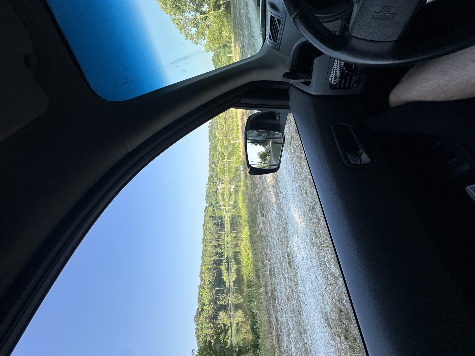 An interior shot from the passenger side of a vehicle shows a view out the window of a body of water surrounded by trees and vegetation under a clear blue sky. The water is calm and reflects the trees. A dirt or gravel road is visible in the foreground. The interior of the car is visible, including part of the dashboard, steering wheel, and door panel. The side mirror reflects a road and trees. The scene appears to be set in a rural or natural area outside of Makiv, Ukraine.